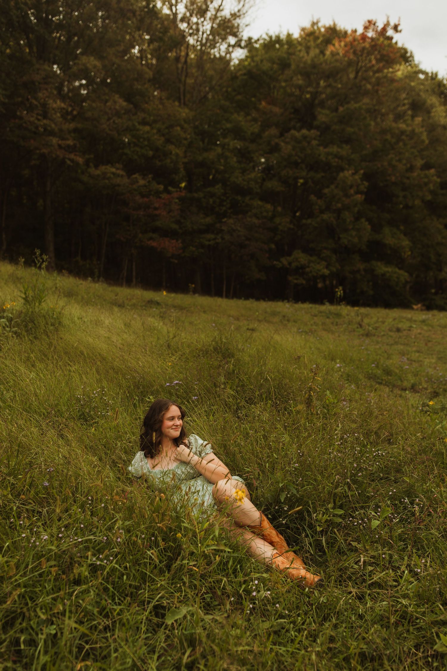 Figure reclines in a summery meadow wearing a floral dress and boots at golden hour.