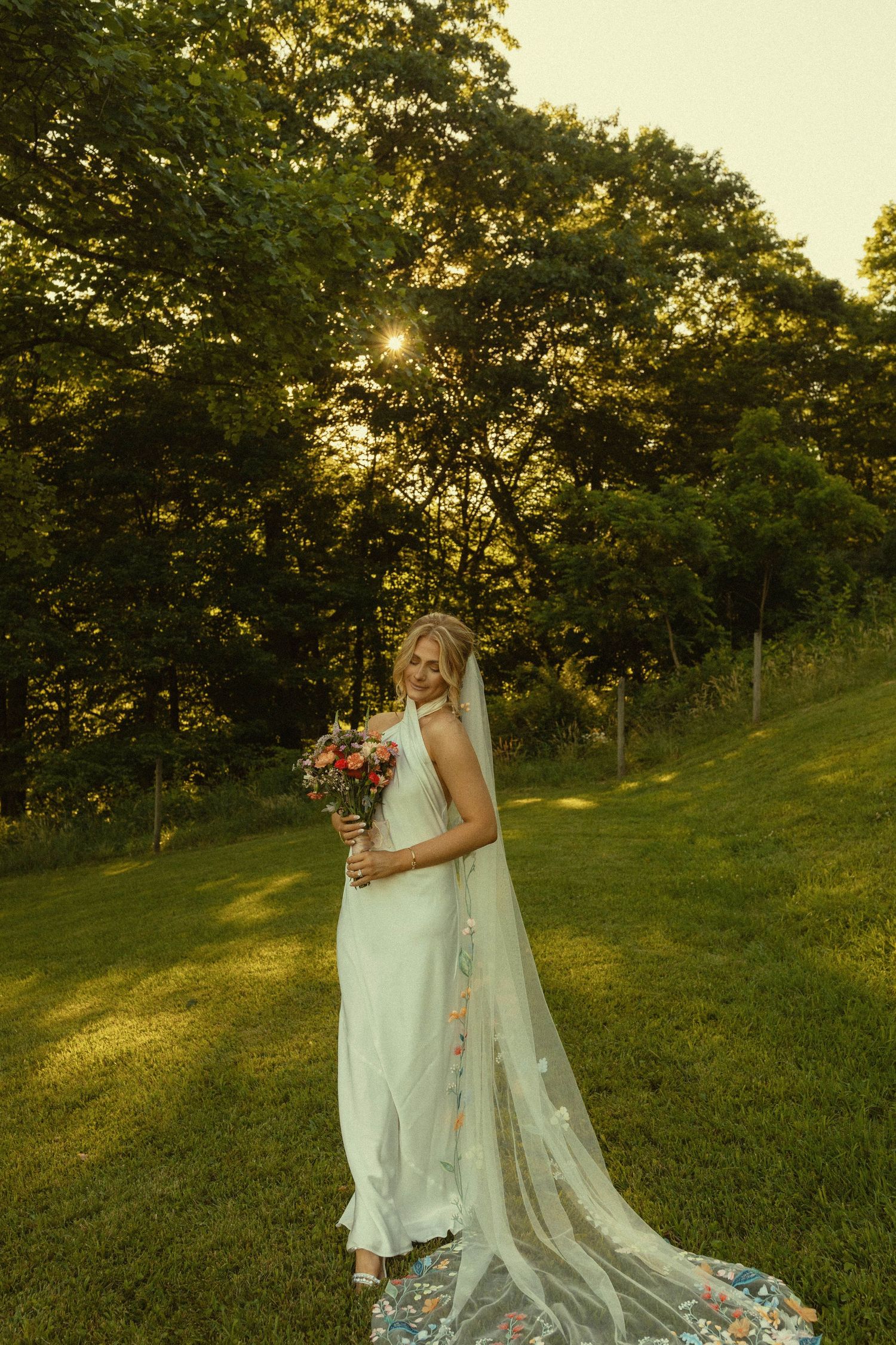 Bride in flowing white dress and veil holds bouquet while standing on grassy hill at sunset.