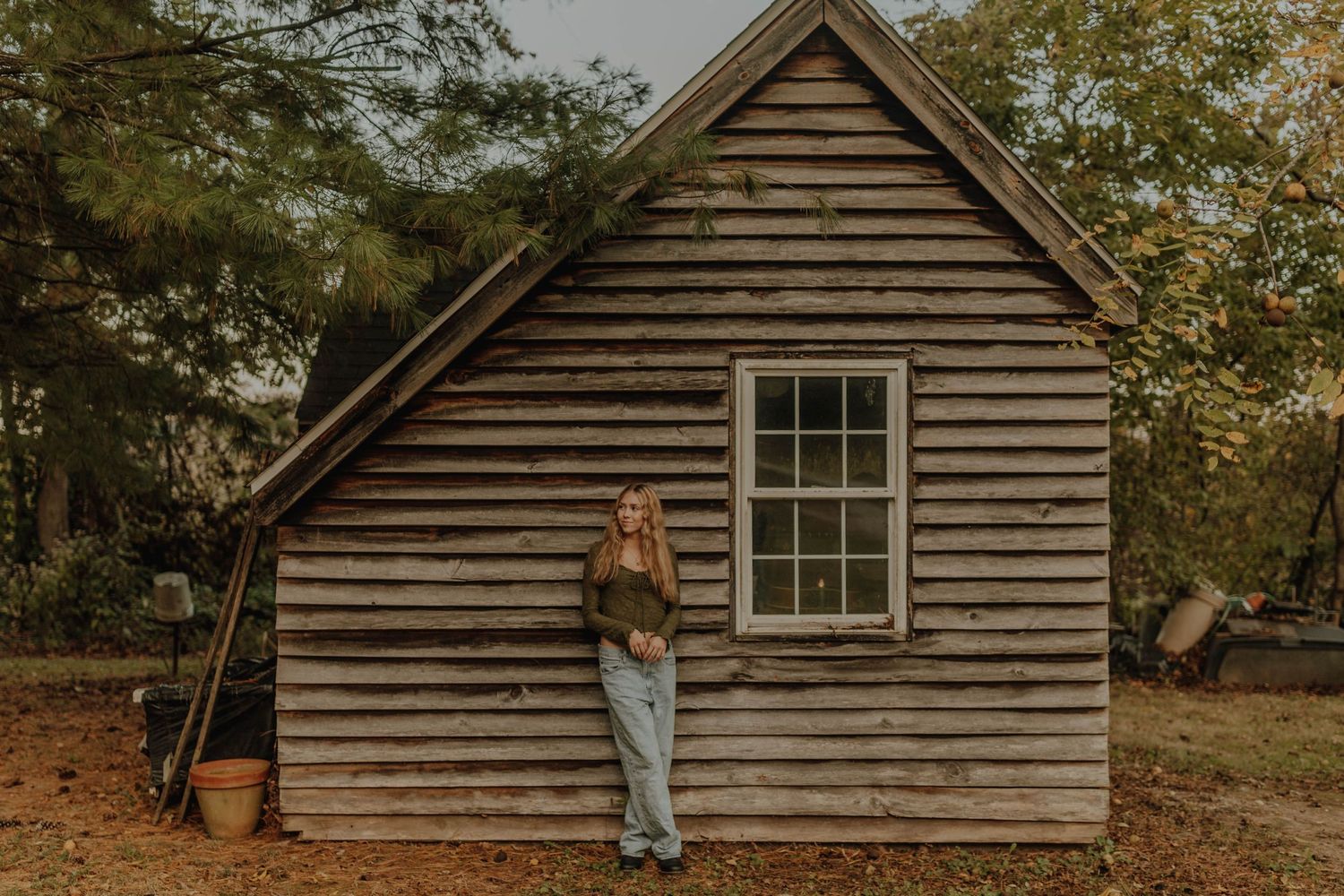 A person leans against a rustic wooden cabin in an autumn setting.