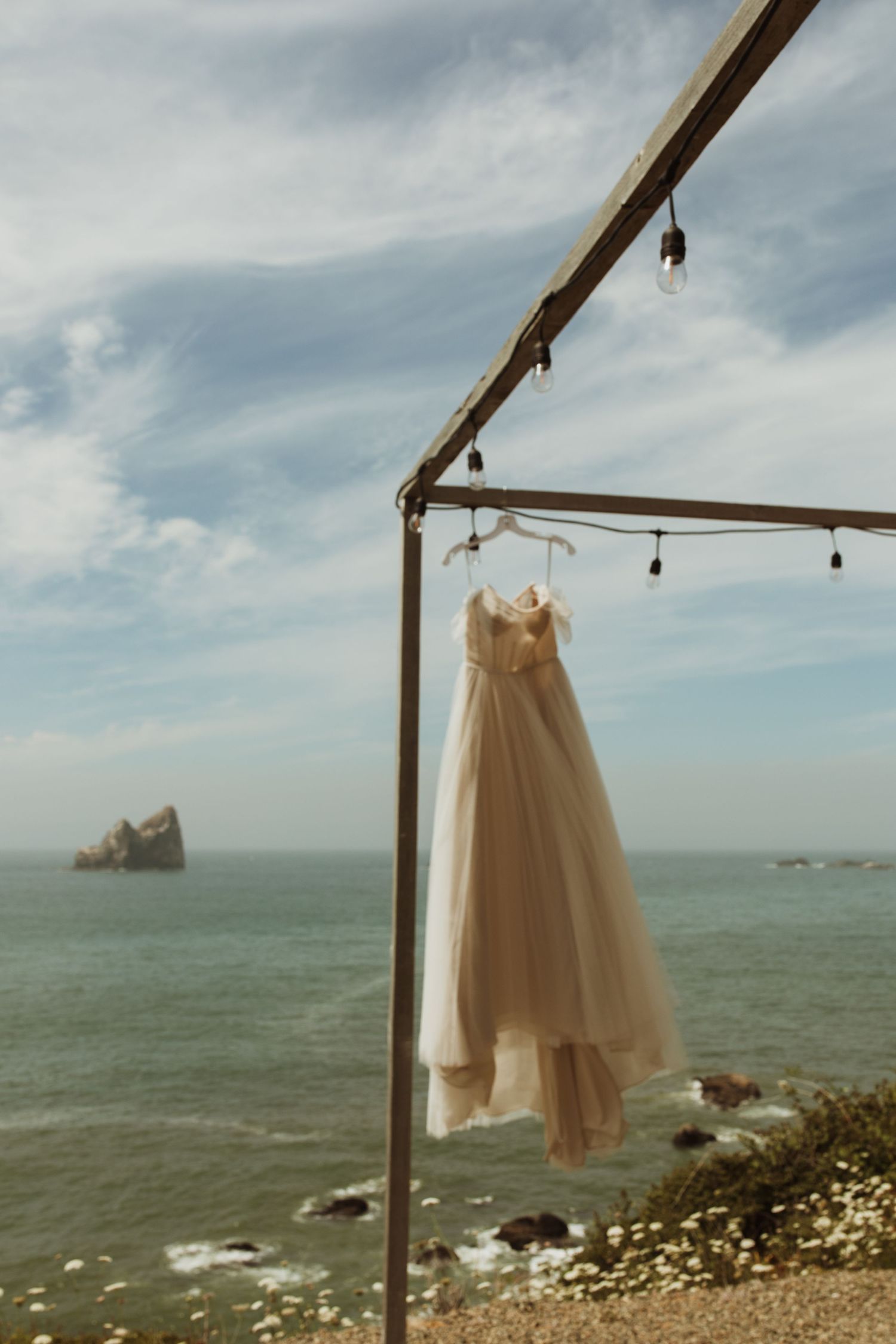 A white wedding dress hangs from string lights with ocean cliffs and rock formations visible in background.