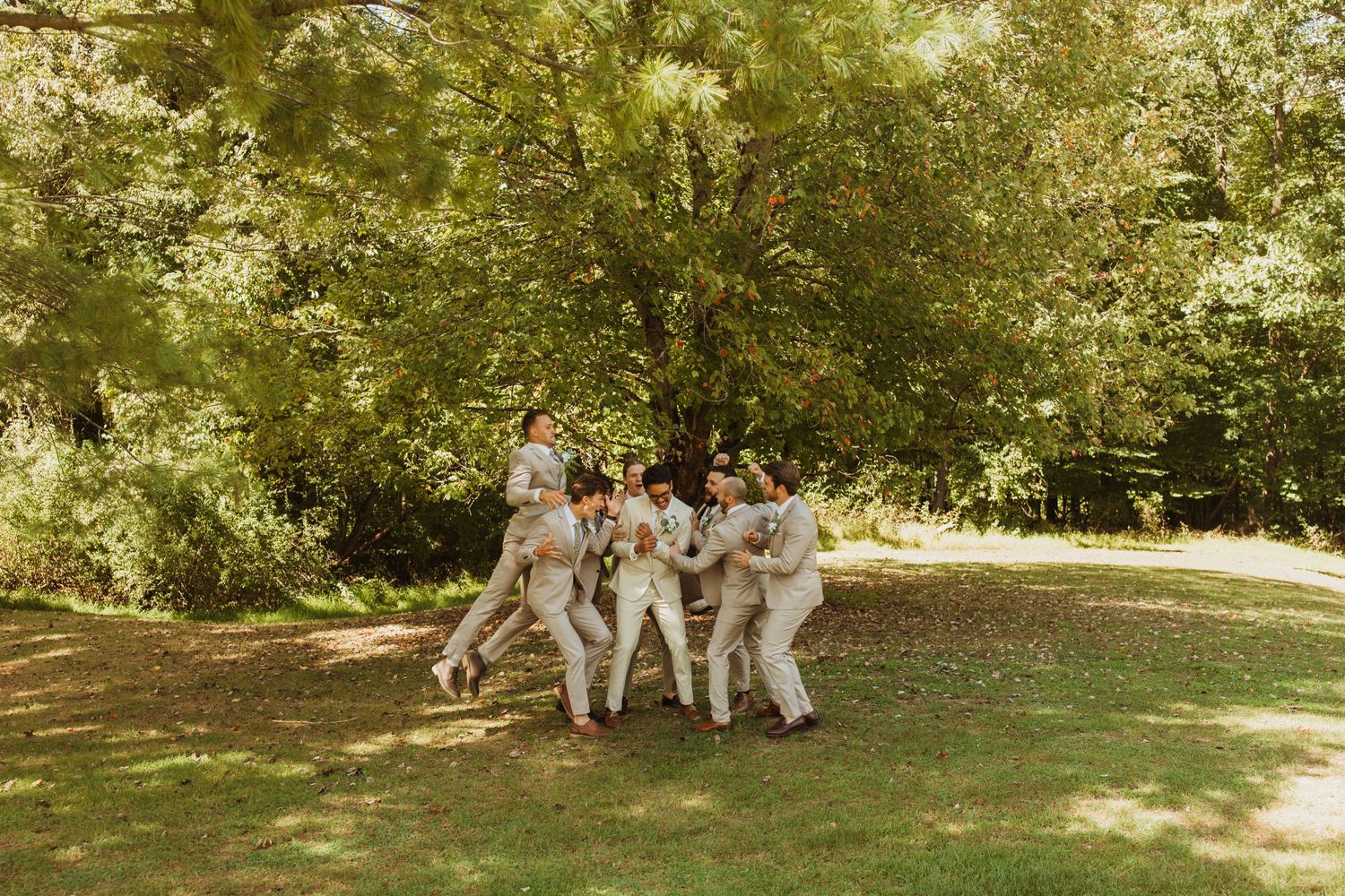Groomsmen in light colored suits playfully pose together on a sunny lawn.