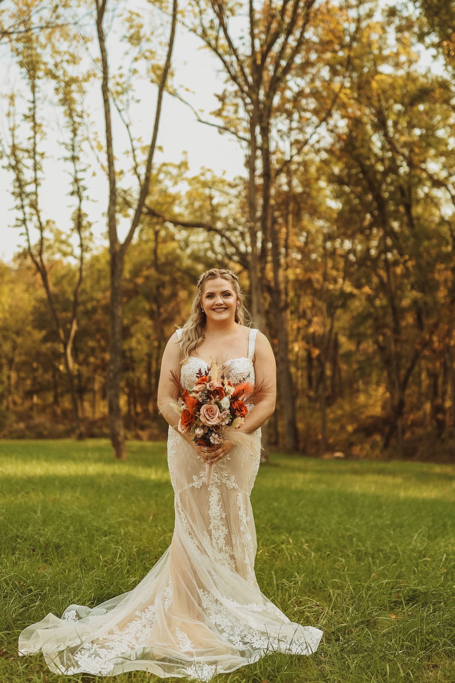 A bride in a lace wedding gown holds an autumn bouquet while standing in a sunlit field.