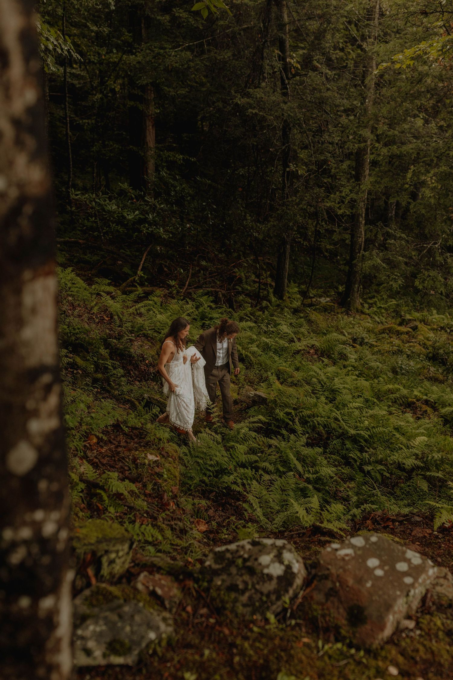 Wedding couple explores a mossy forest hillside in elegant attire.