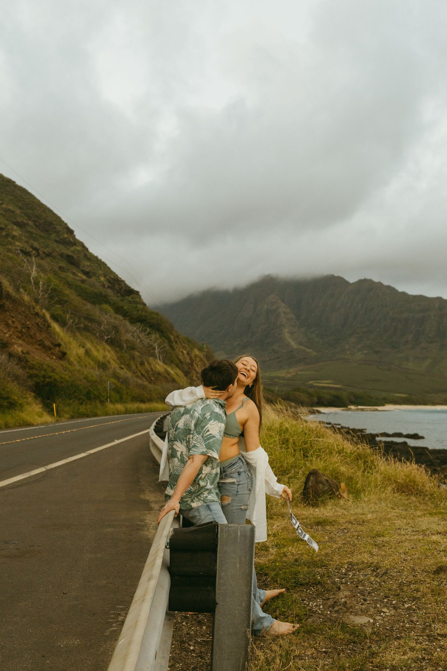 Playful pair posing on a guardrail along a coastal mountain road during overcast weather.