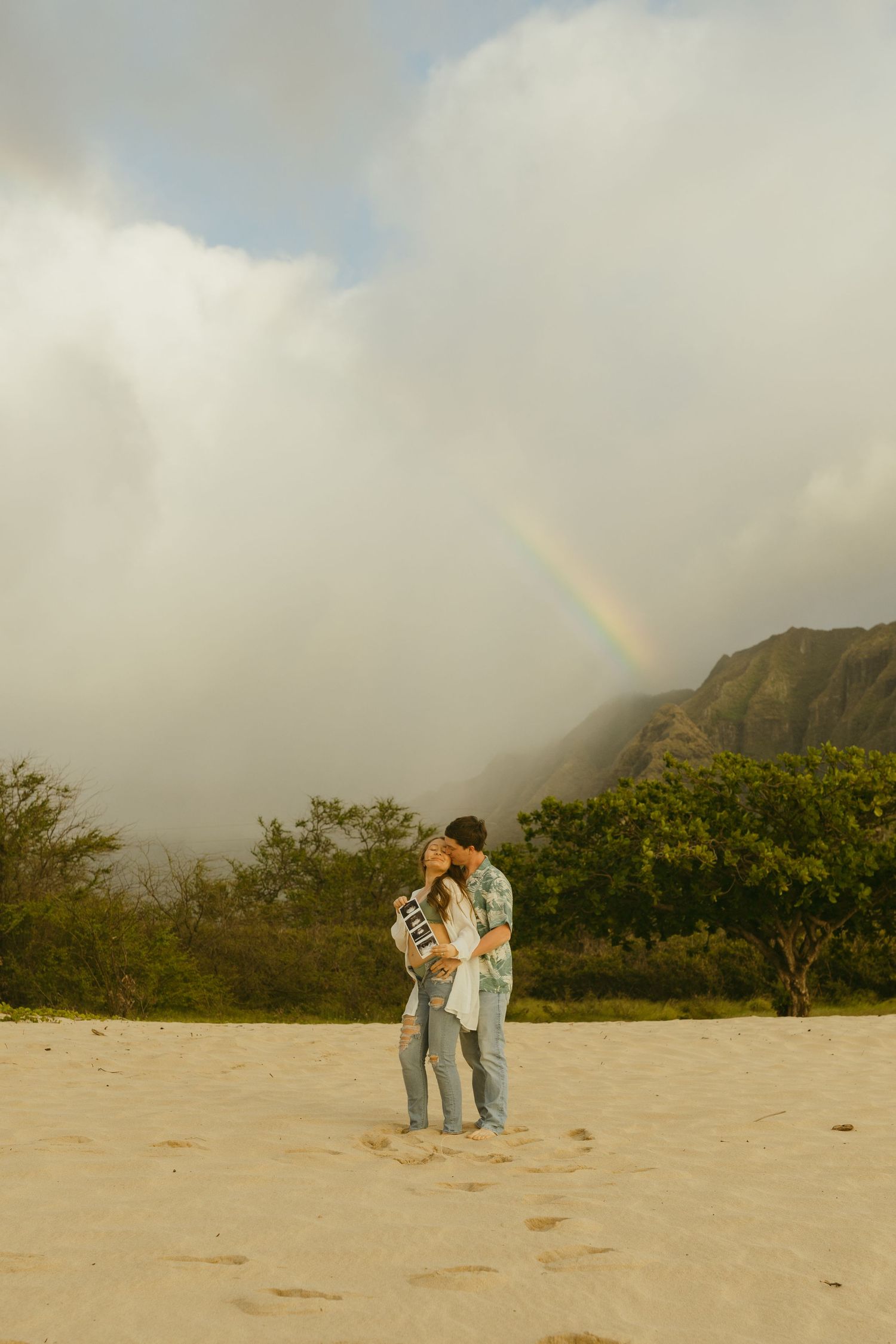 Couple embraces on sandy beach with rainbow and mountains in background.