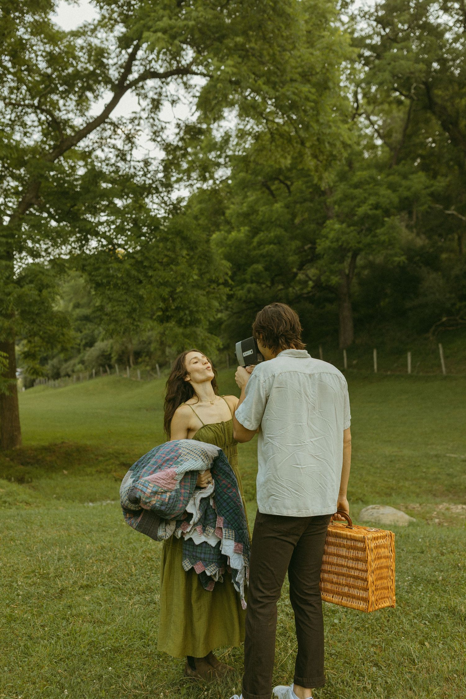 A couple shares laughter while holding picnic supplies under a tree in a park.