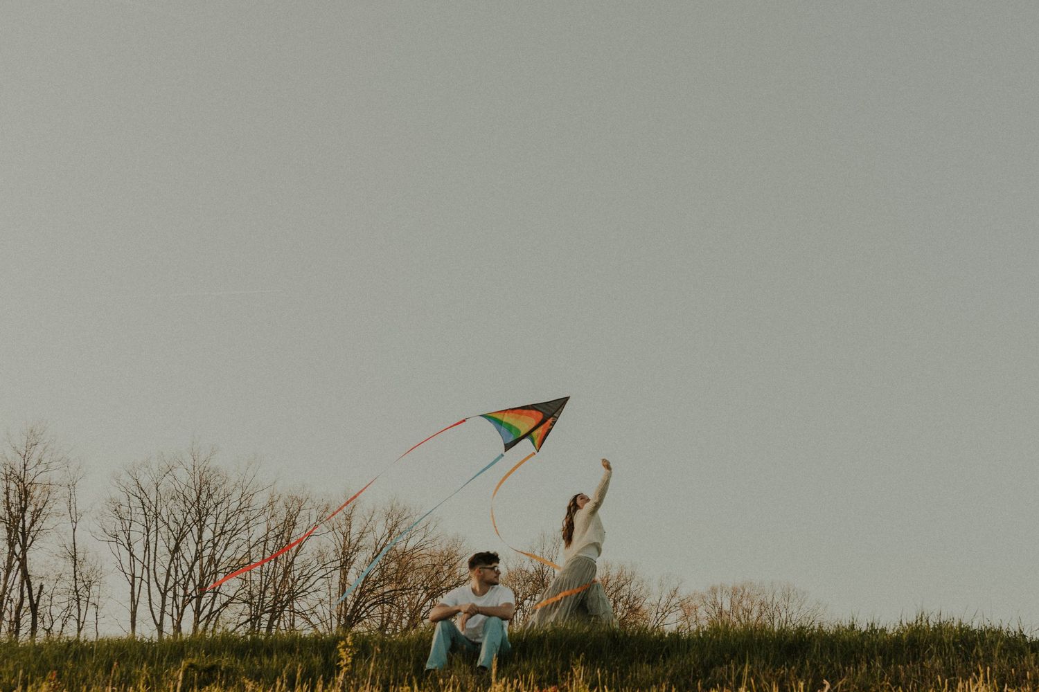 A couple flies a colorful kite on a hilltop against a cloudy sky during golden hour.