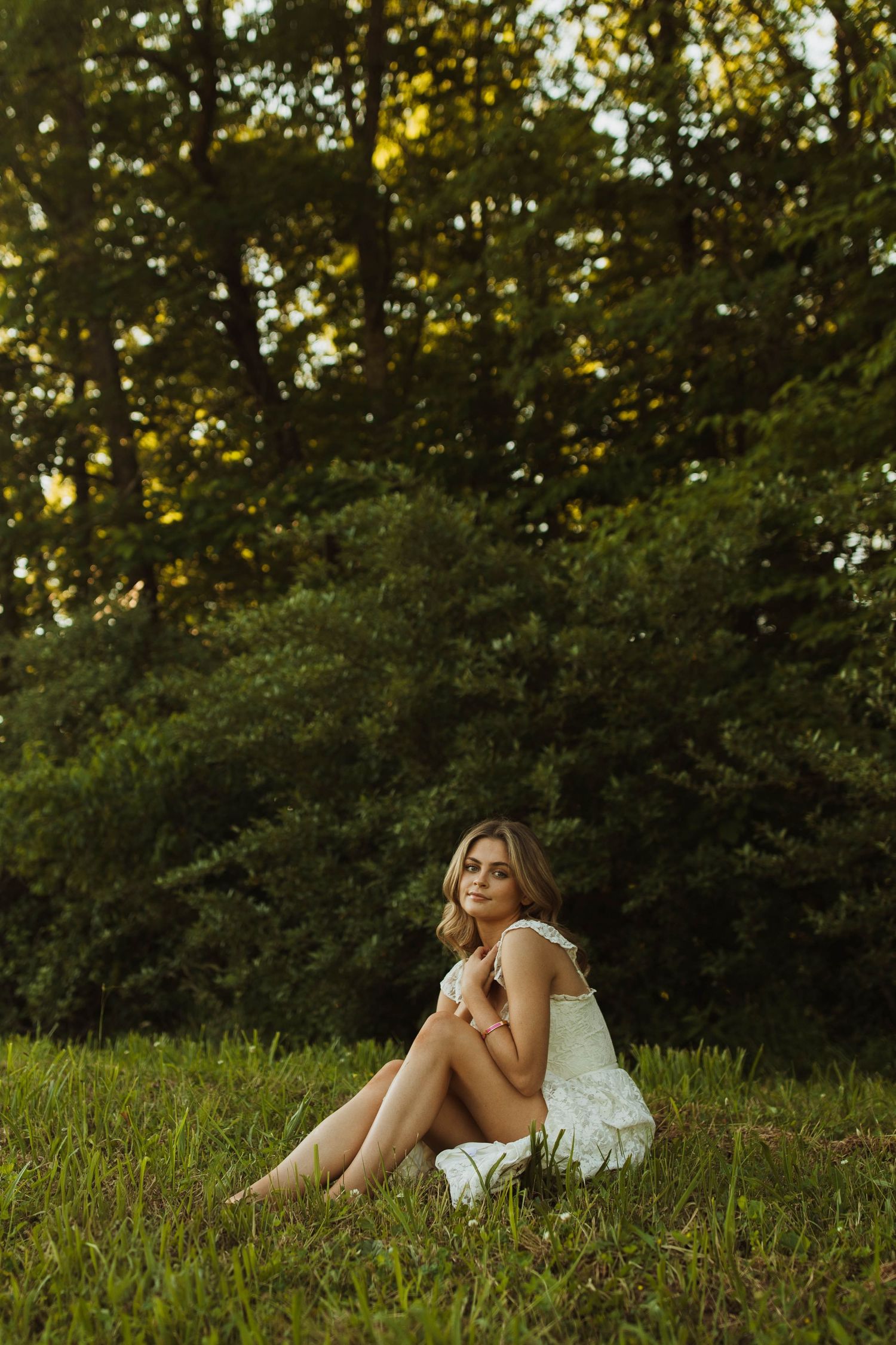 A woman in a white sundress sits on lush grass at golden hour with trees in the background.