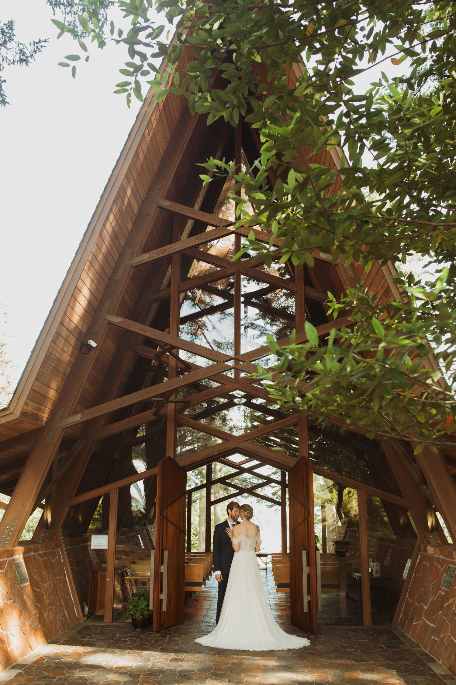 Wedding couple poses under dramatic wooden A-frame chapel architecture surrounded by greenery.