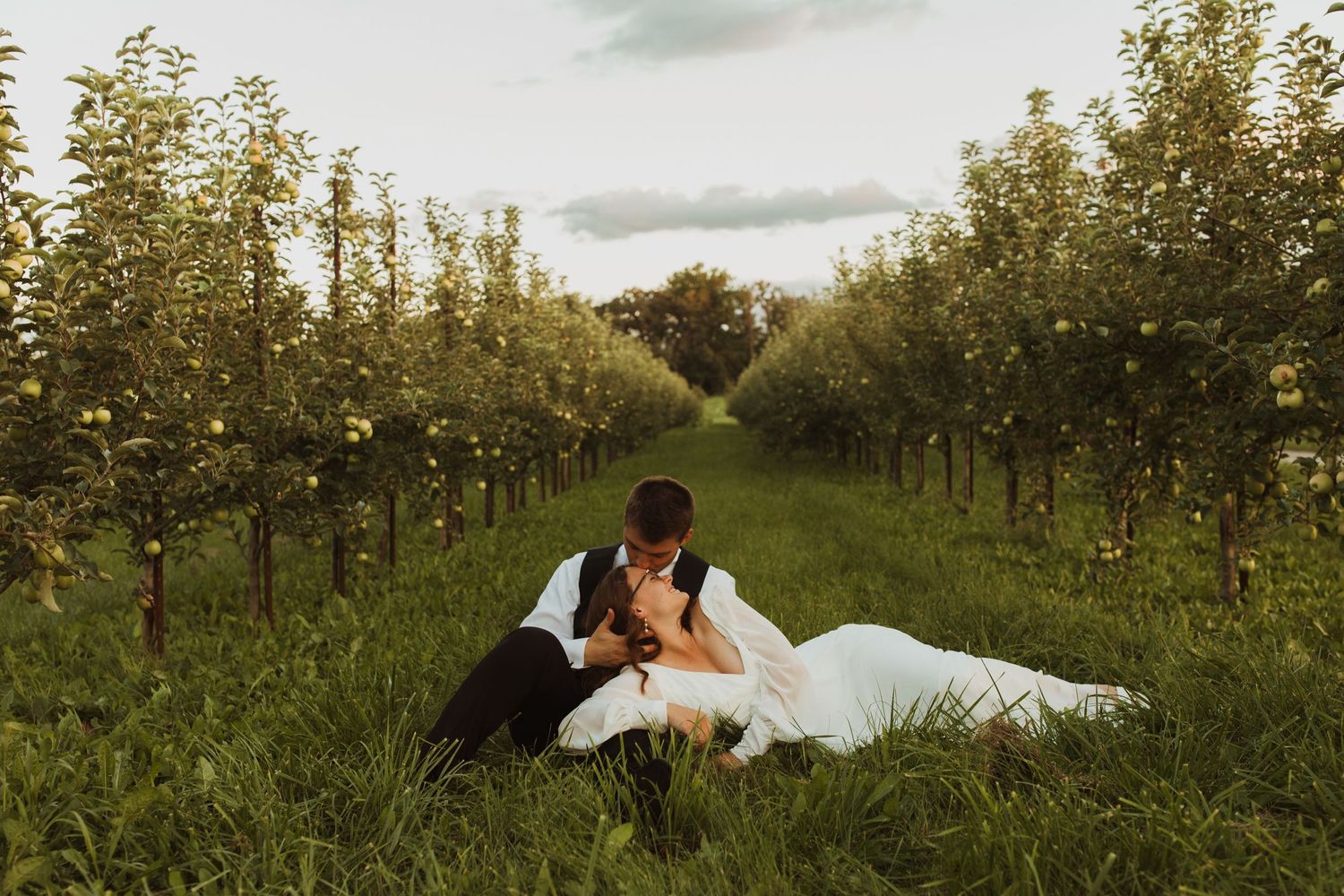A romantic wedding couple lying in a scenic apple orchard during sunset.