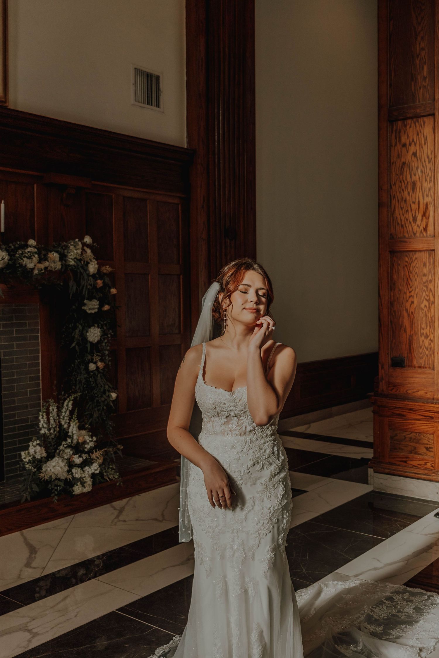 Bride in lace gown poses elegantly beside floral arrangements in historic venue.