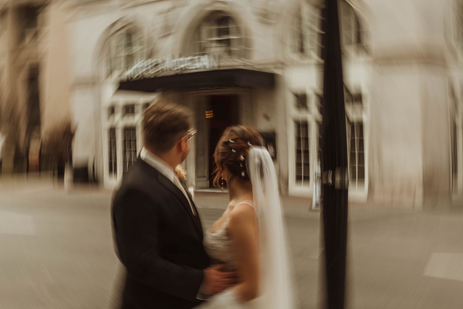 A romantic wedding couple poses outside an elegant venue entrance with a soft sepia filter effect.