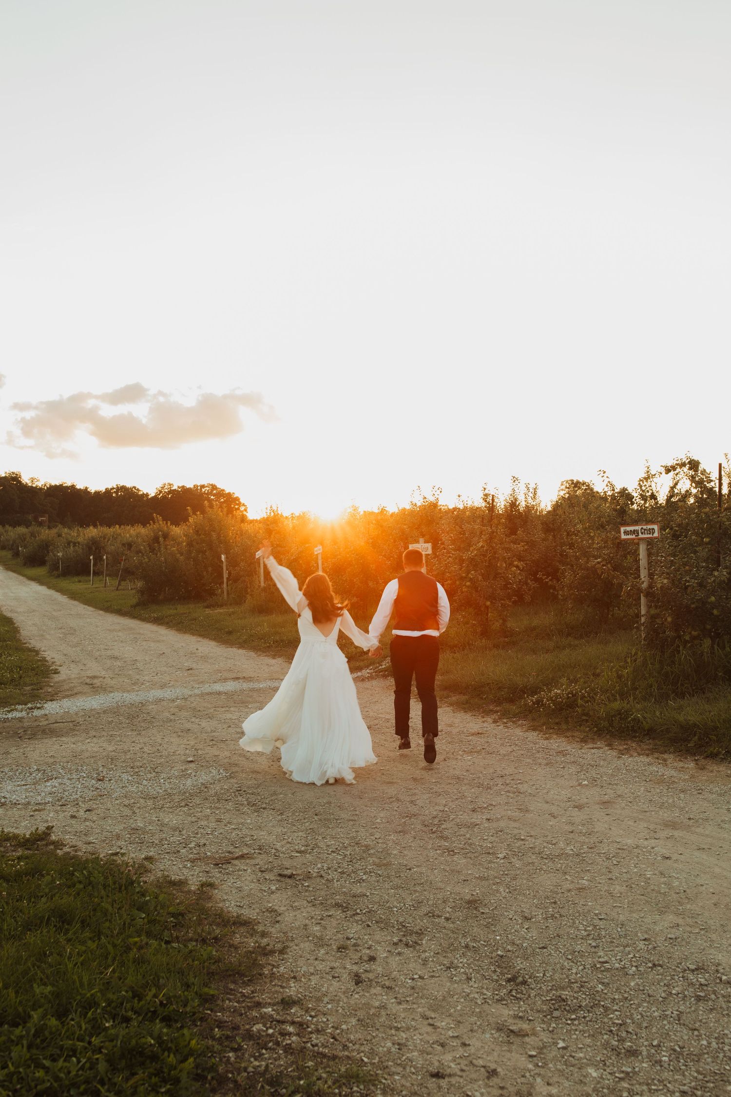 Newlyweds running hand in hand down a dirt path at sunset.