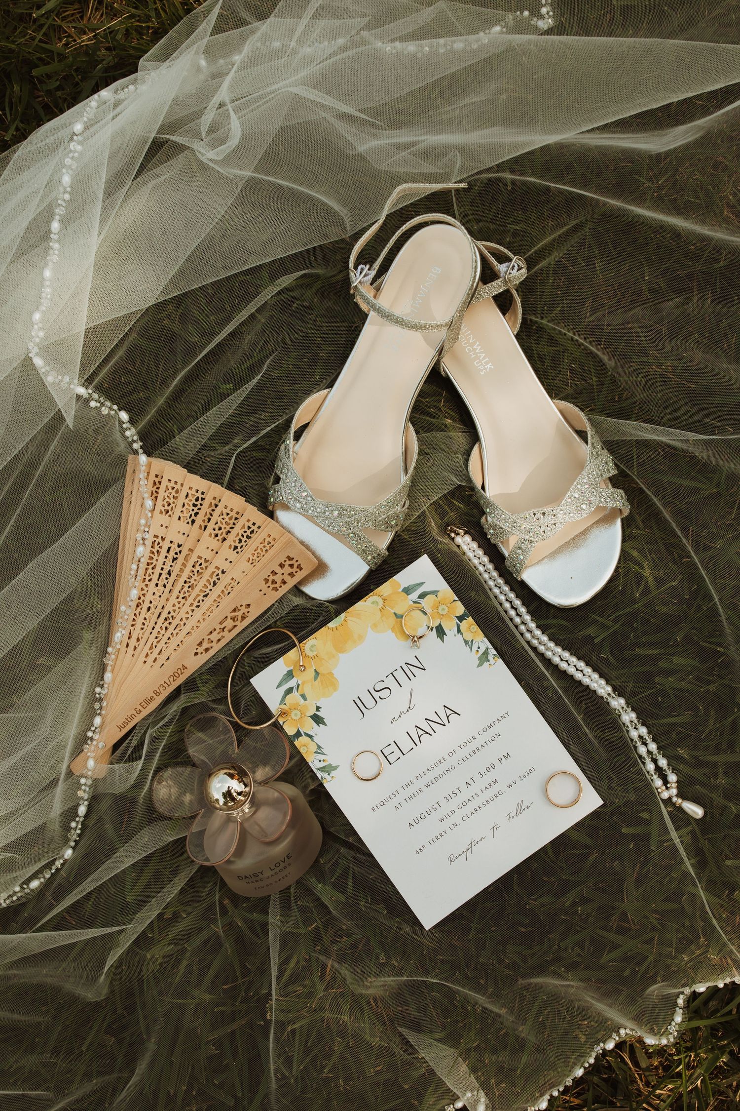 Wedding accessories including silver sandals, a decorative fan, invitation card and veil arranged on dark fabric.