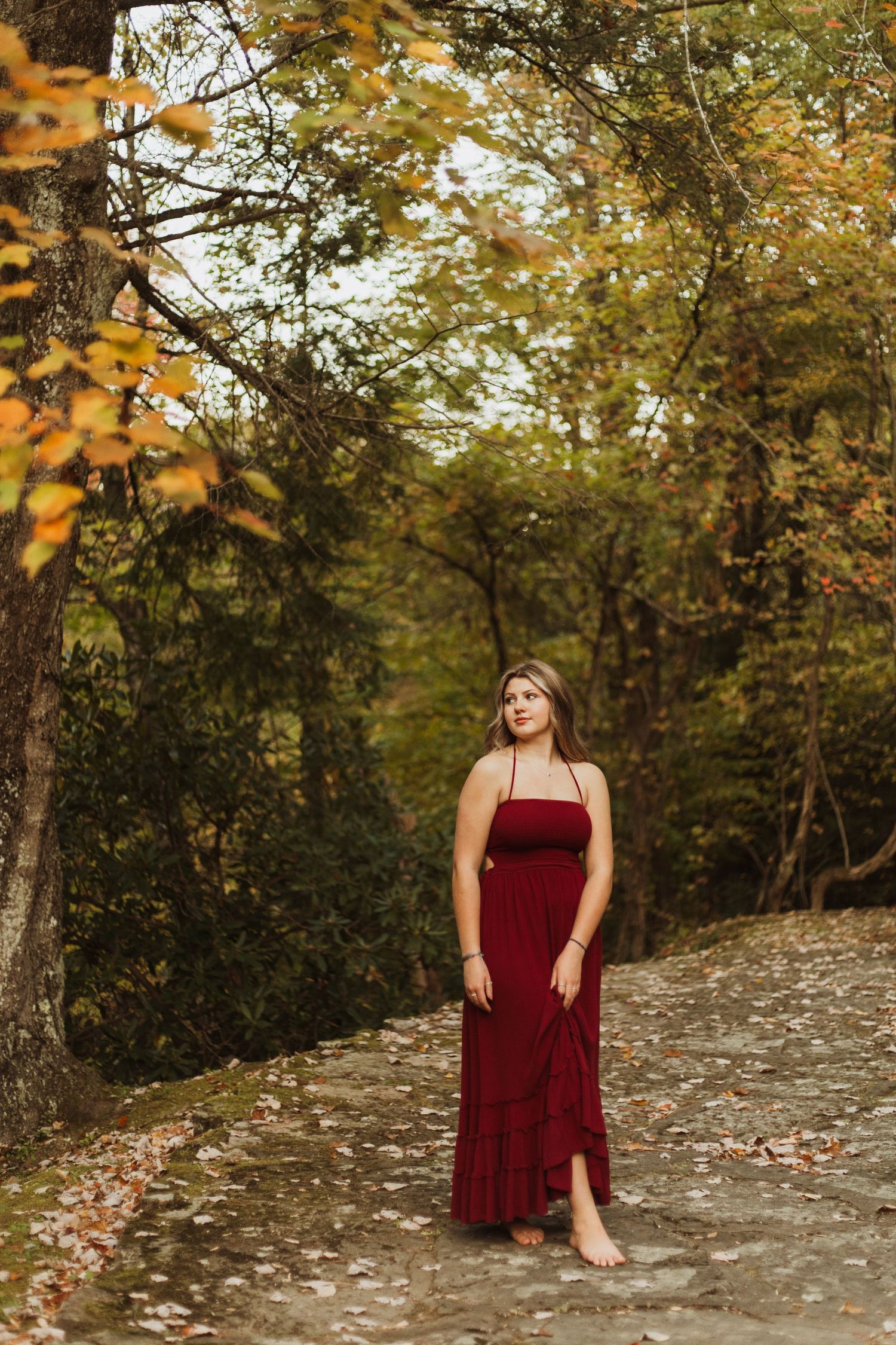 A burgundy maxi dress stands out along a forest path surrounded by golden fall foliage.