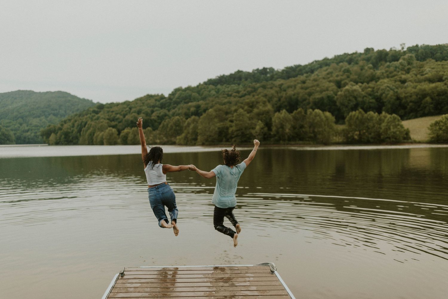 Two figures leaping joyfully off a wooden dock into a calm lake surrounded by hills.