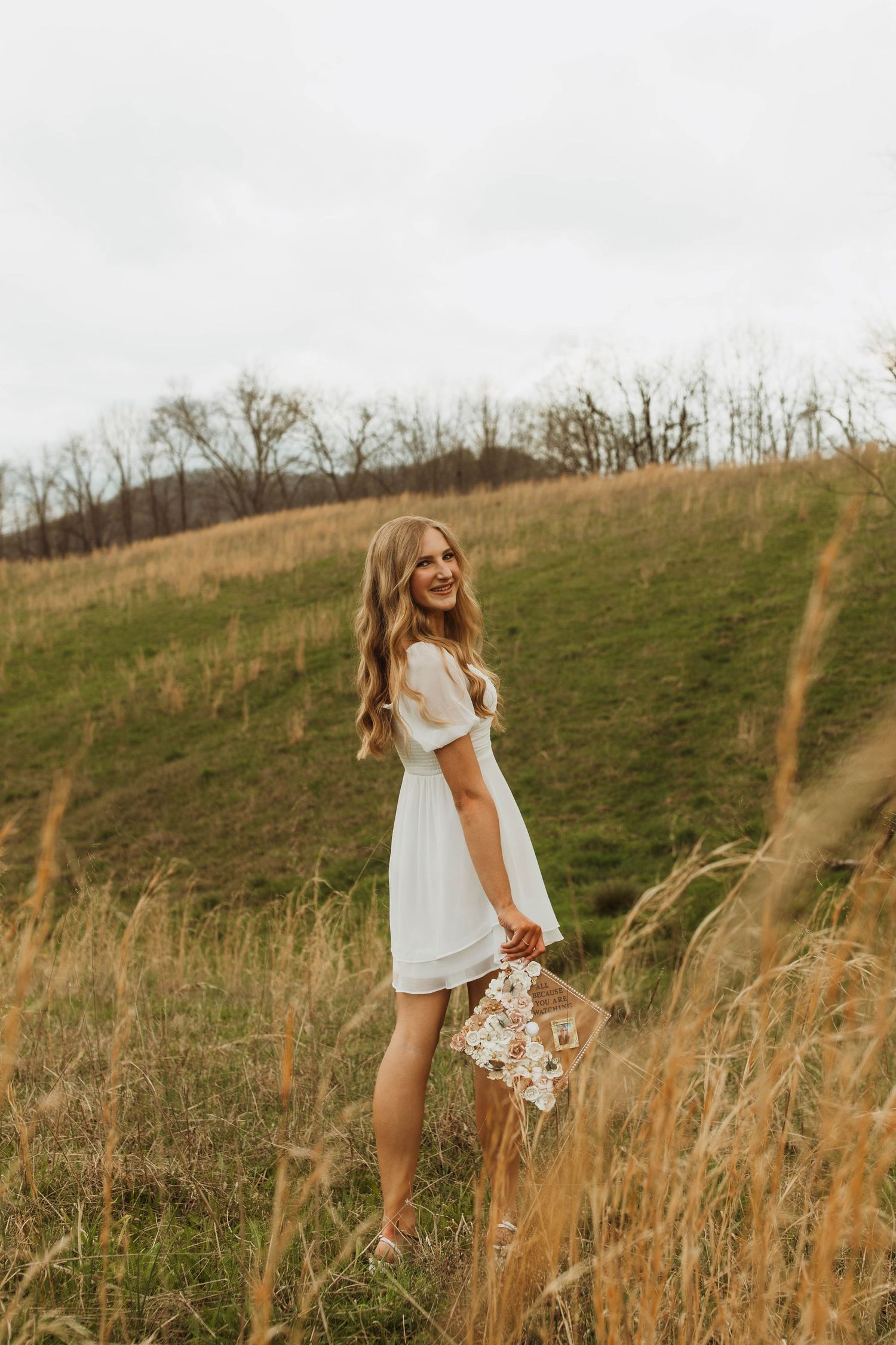 Someone stands in a field of tall grass holding flowers while wearing a white summer dress.