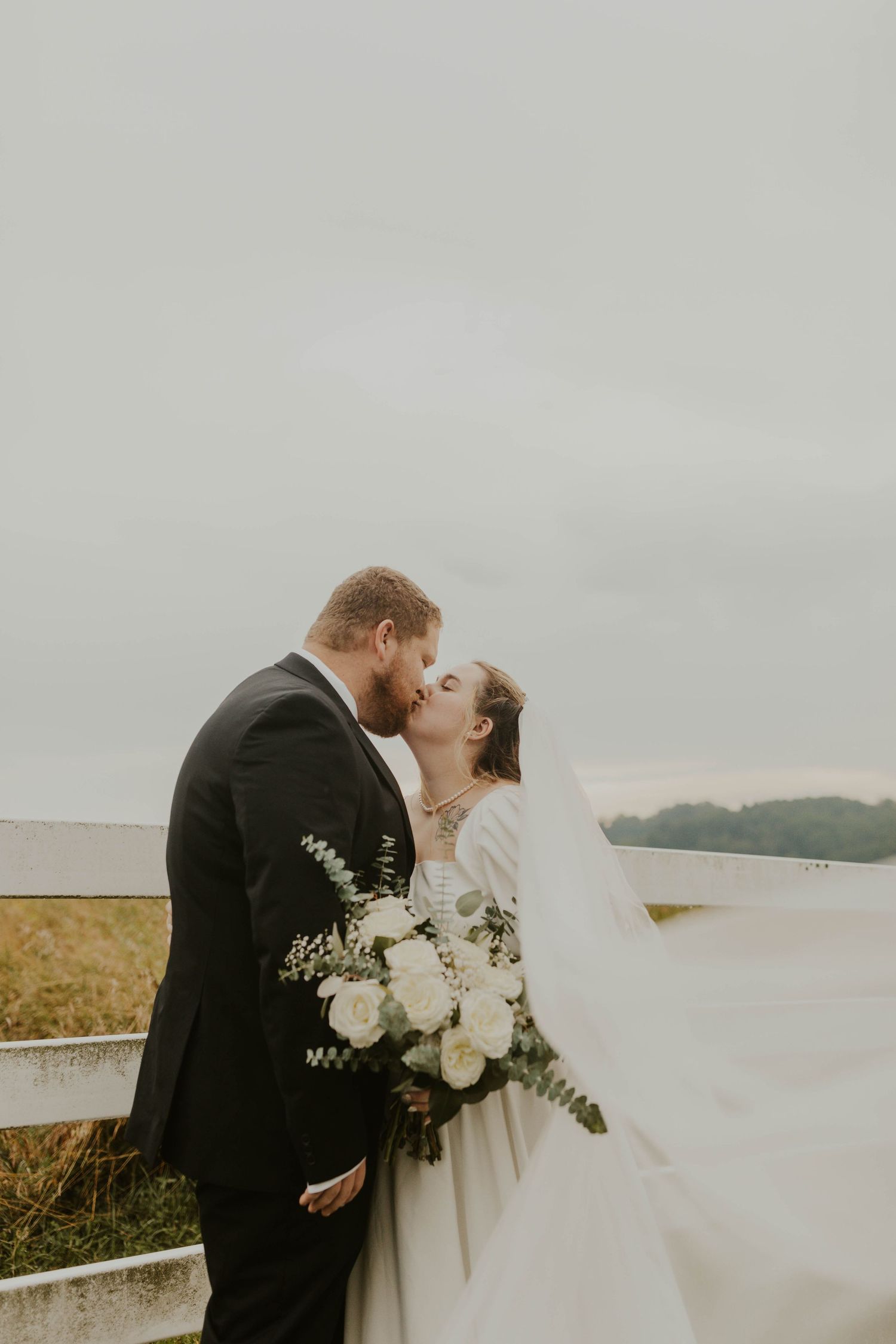 Romantic wedding couple embracing with white bouquet against cloudy sky.