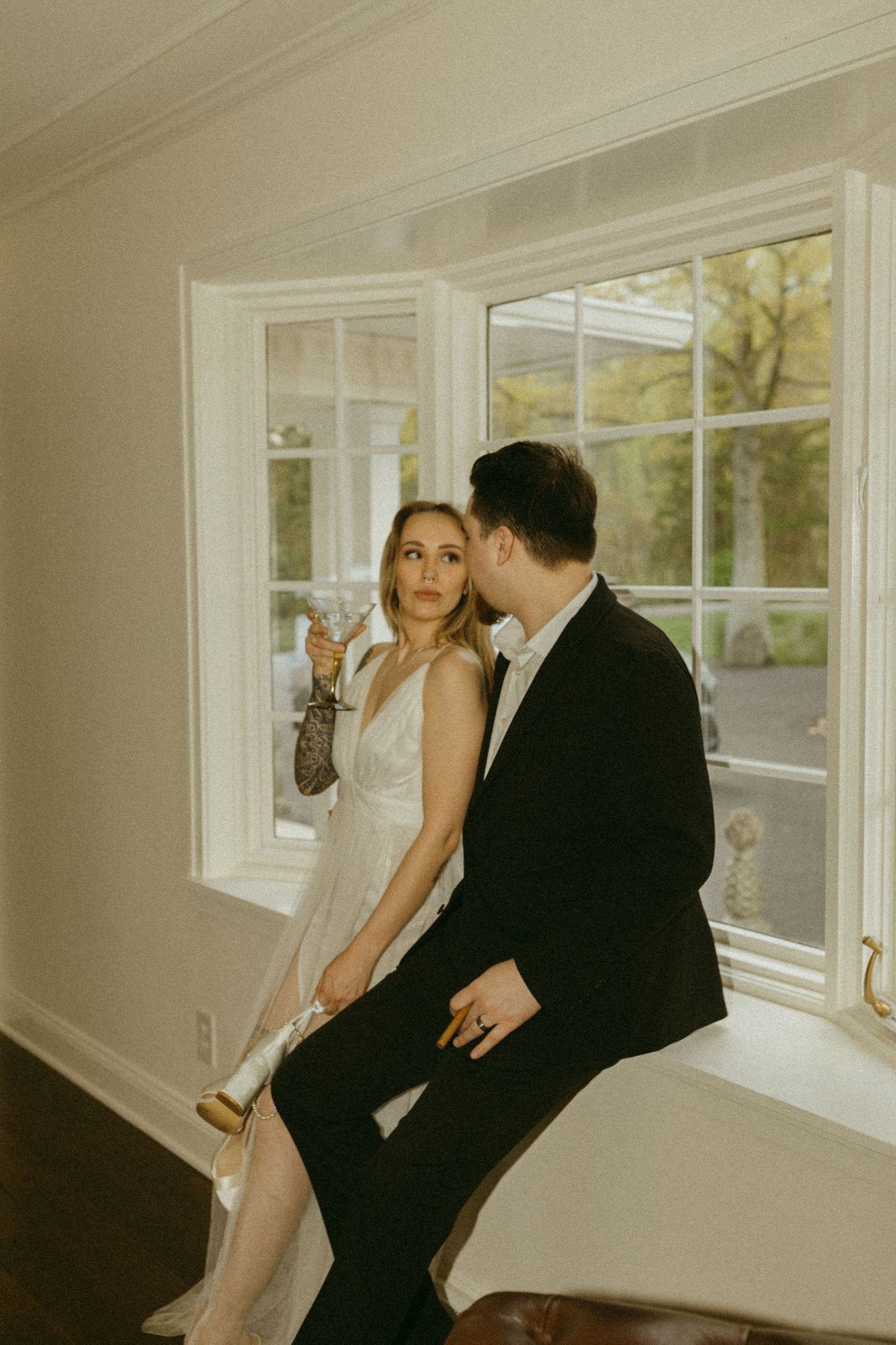 Elegant couple enjoying champagne while seated on a windowsill in formal wear.