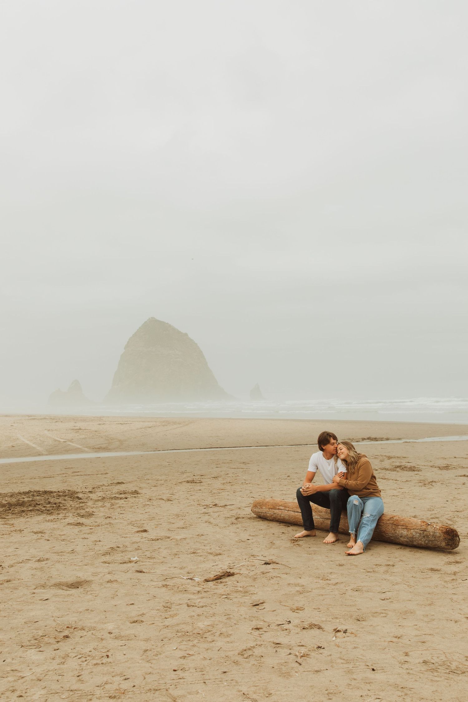 Couple sitting on driftwood log at foggy Cannon Beach with Haystack Rock visible.