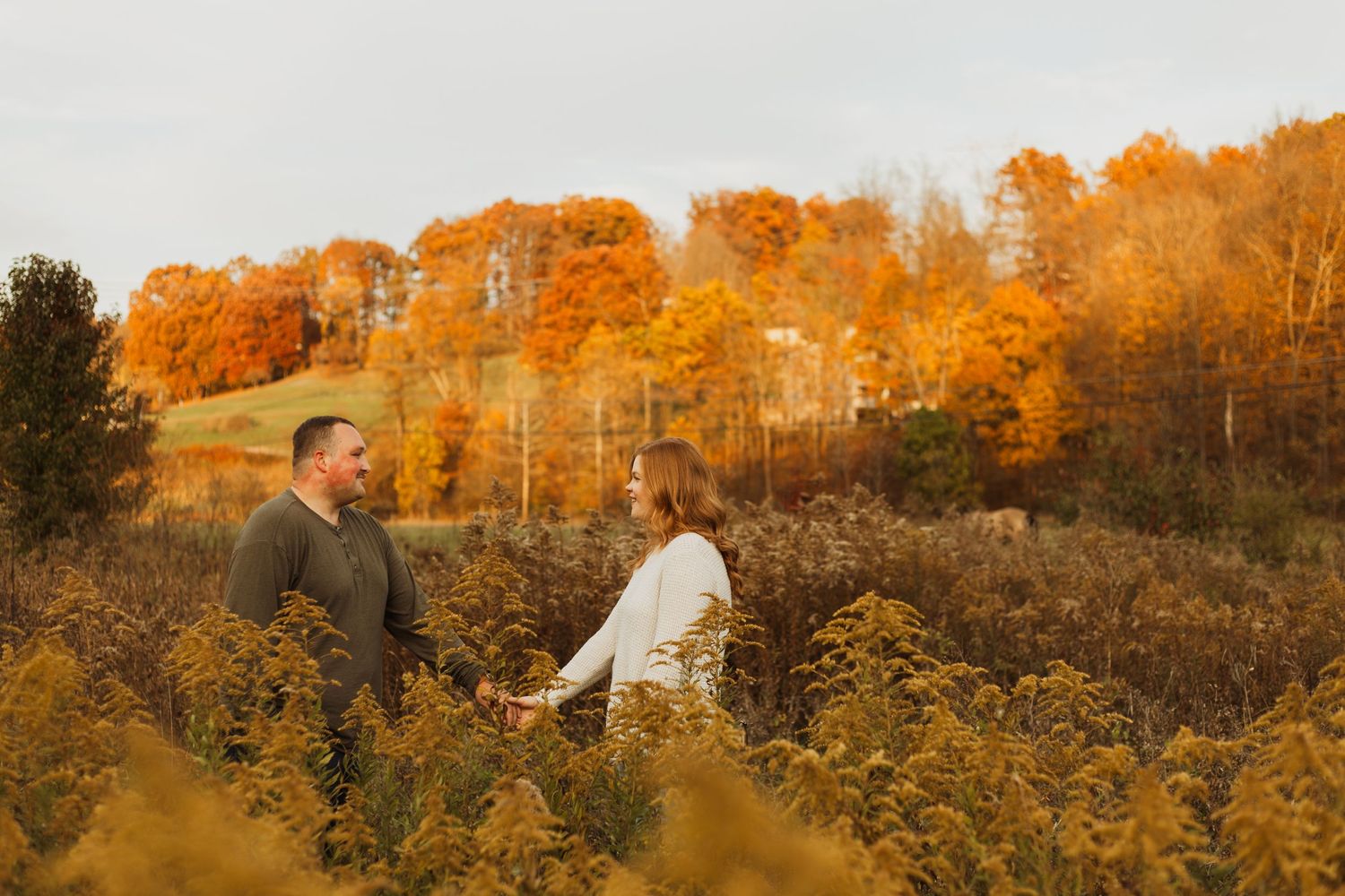 A couple standing in tall grass with vibrant orange fall foliage behind them.