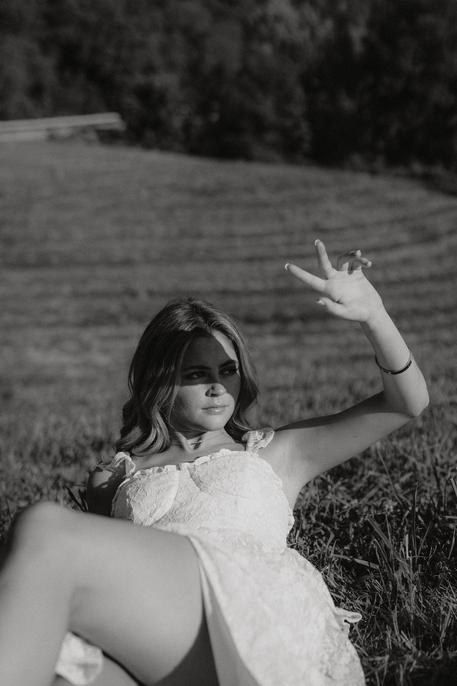 Black and white photo of someone in a white lace dress lying in grass while making a peace sign gesture.