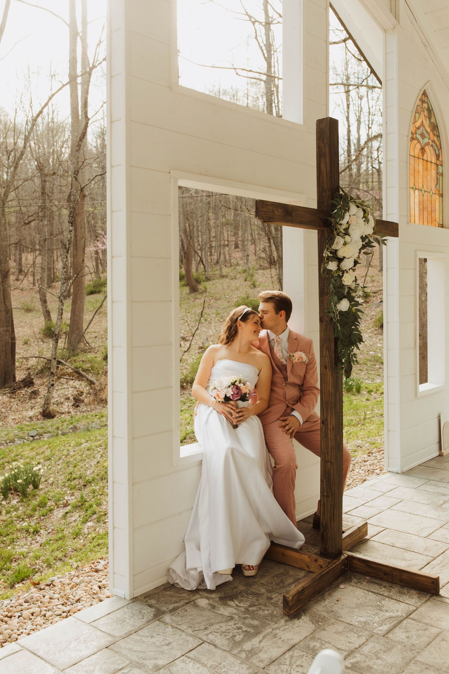 Wedding couple posing by wooden cross at rustic chapel with spring forest background.