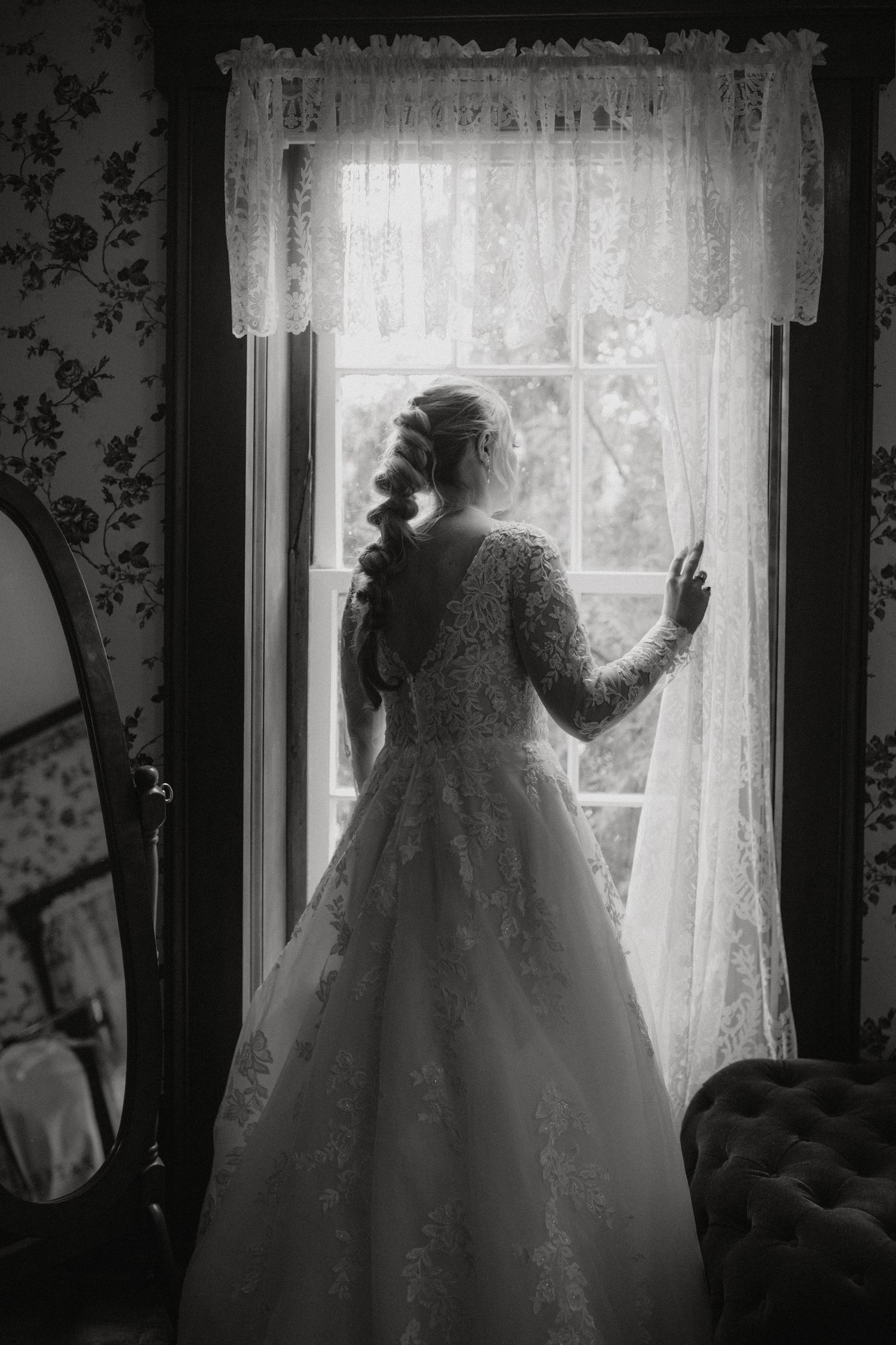 A silhouette of a bride in a lace gown standing by a window with vintage curtains.