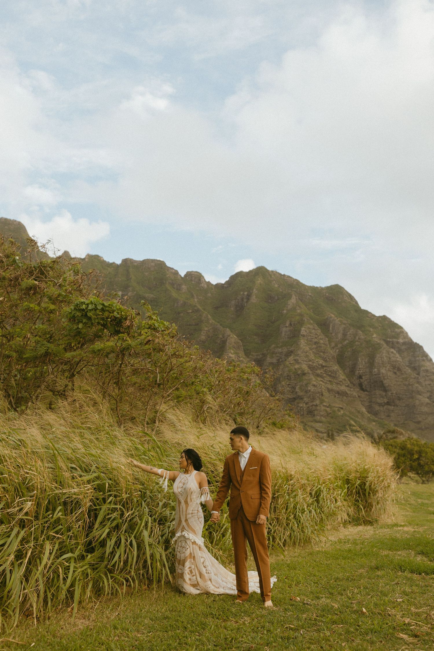 A couple poses in formal attire against a backdrop of majestic mountain ranges and tall grass.
