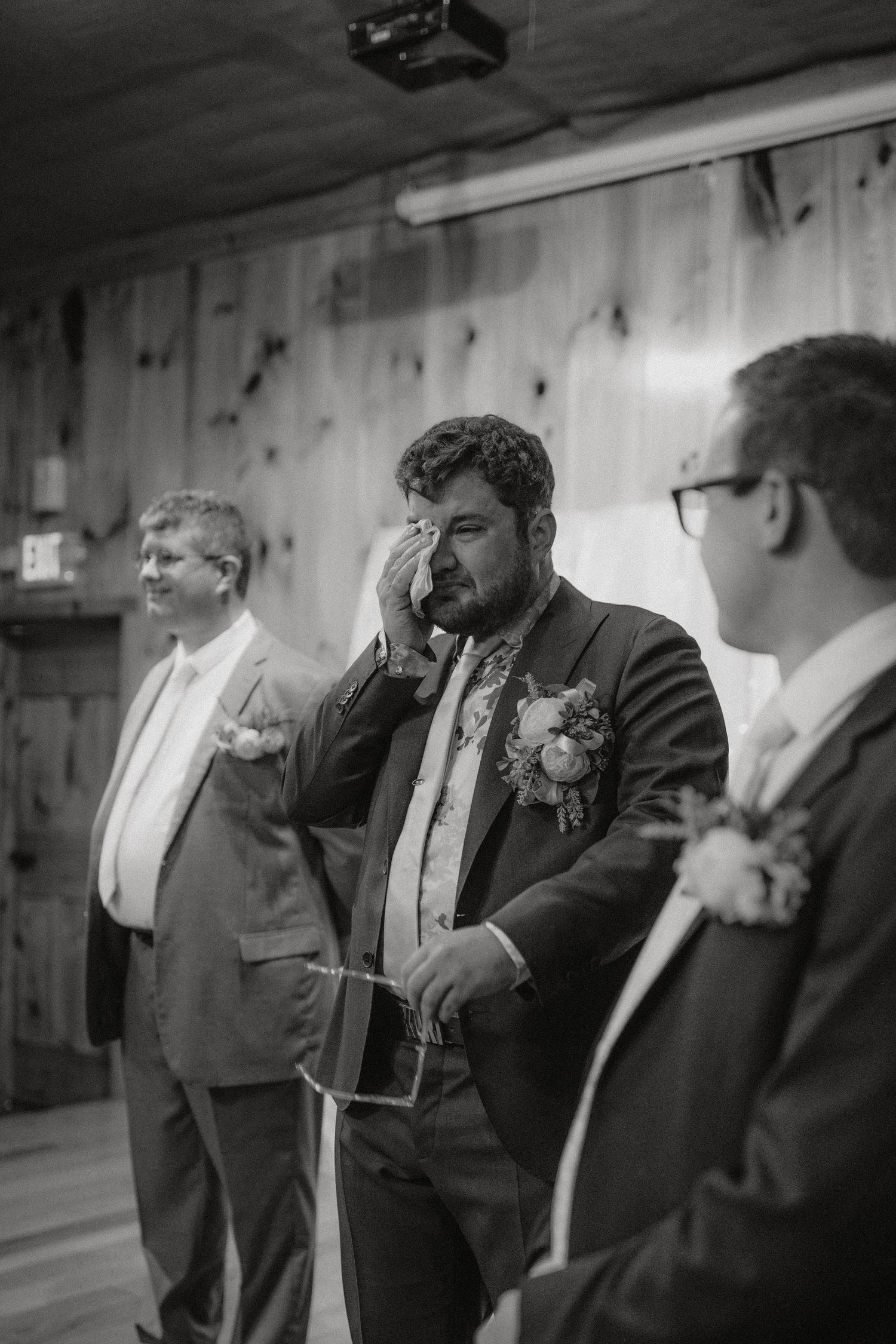Three groomsmen in suits sharing an emotional moment during a wedding ceremony indoors.