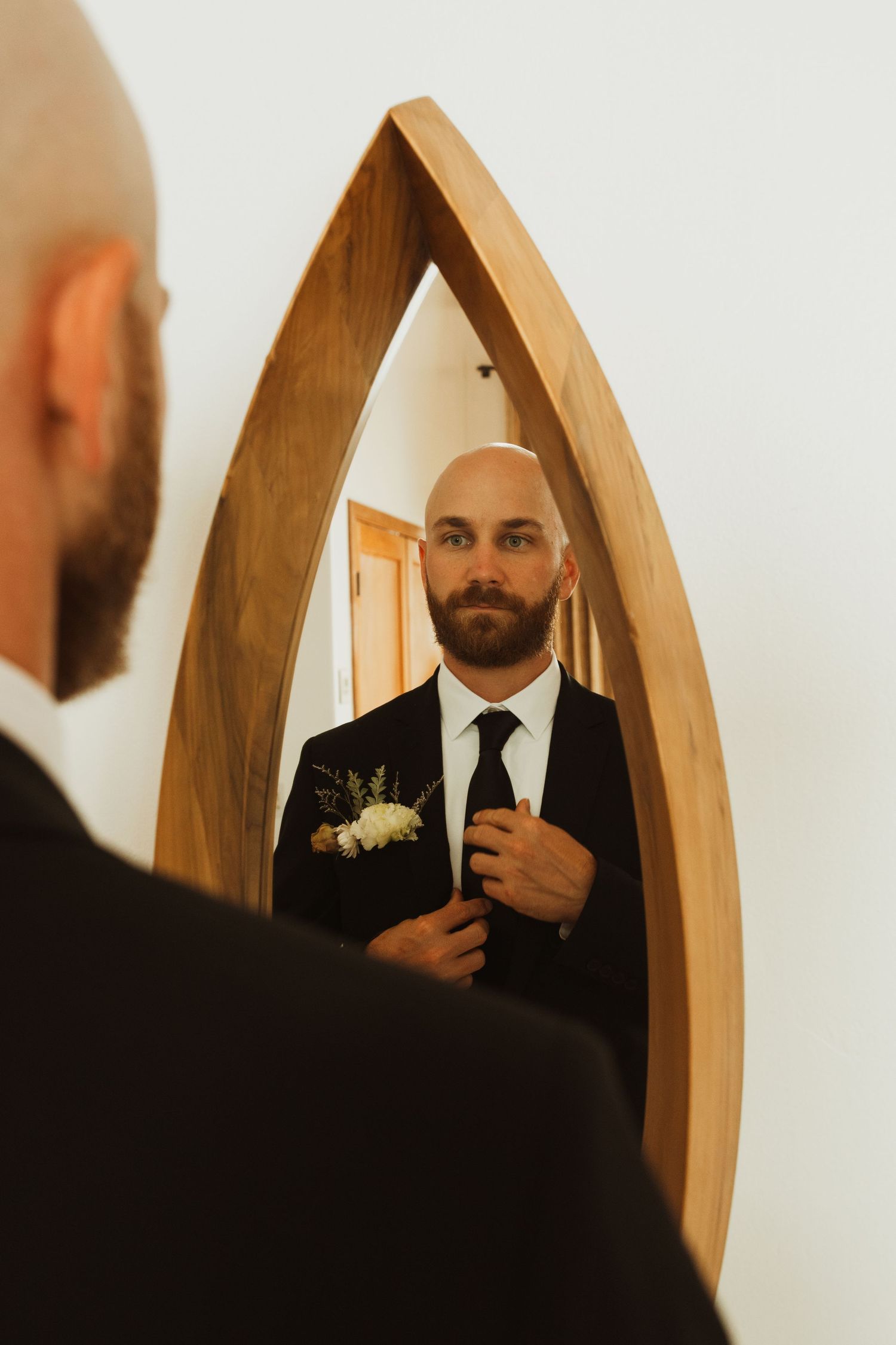Groom adjusting tie while looking in wooden arch-shaped mirror.