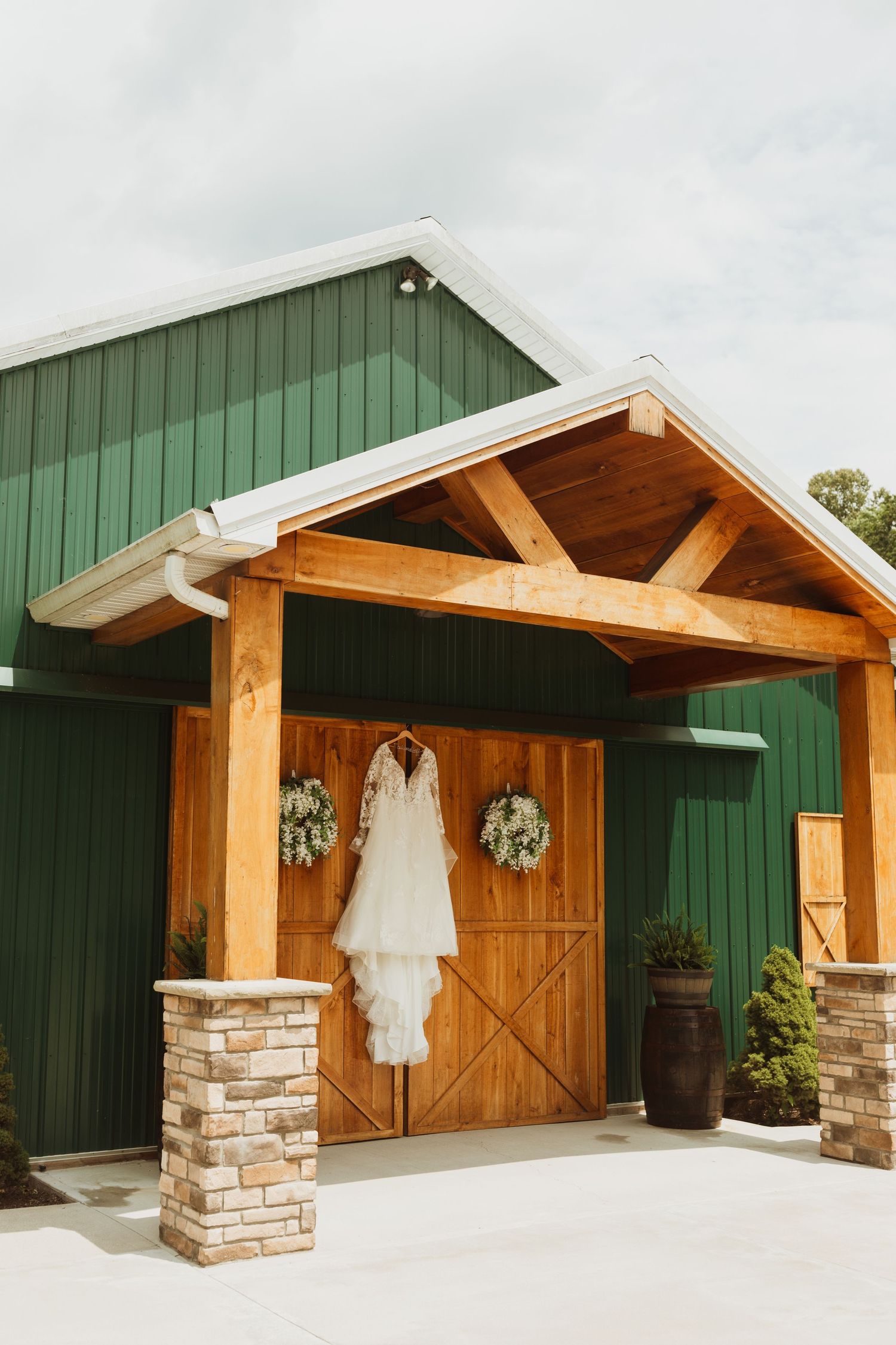 White wedding dress hanging outside rustic green barn venue with wooden beams and stone accents.