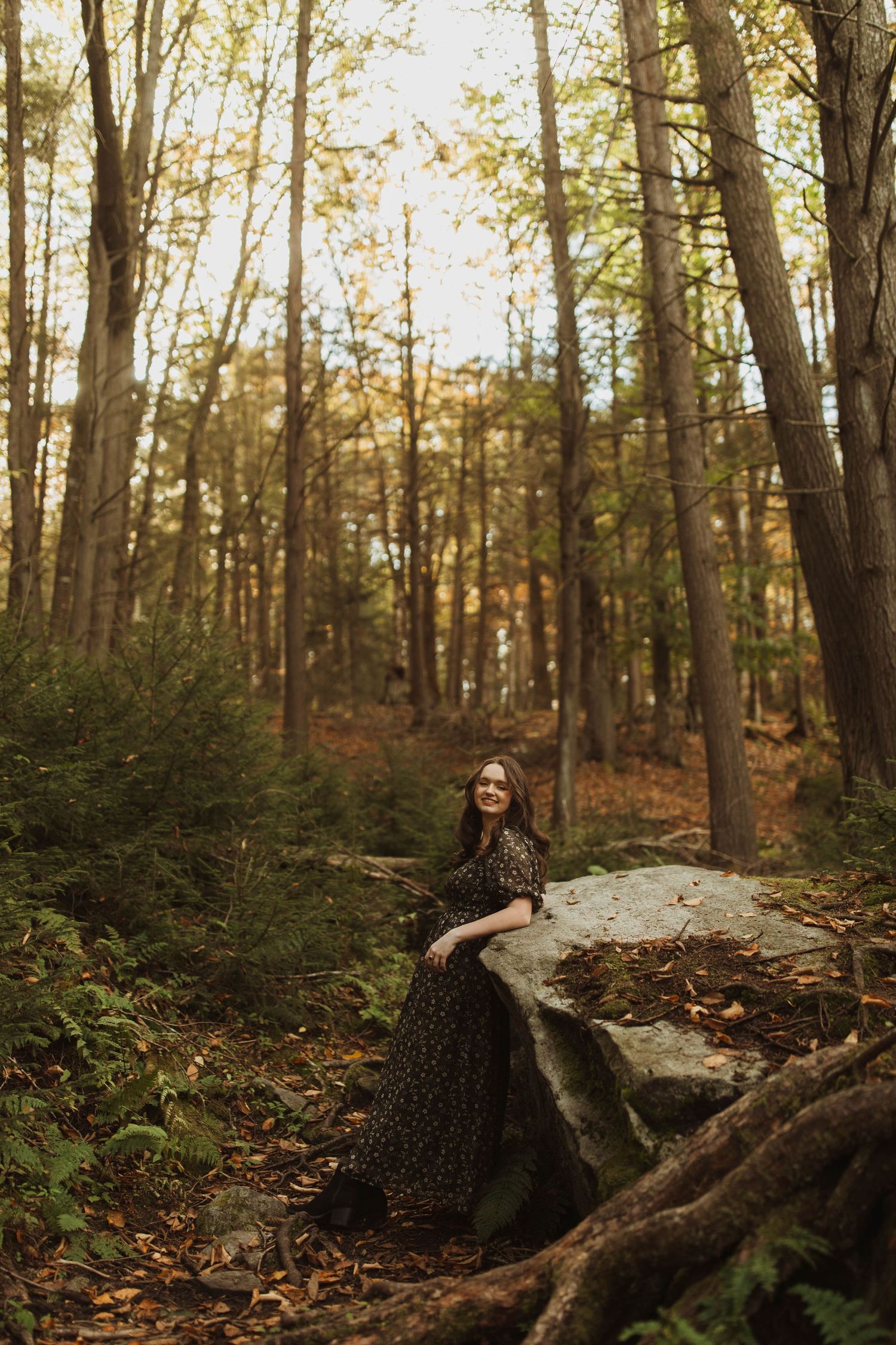 Someone in a dark dress poses on a large rock in a moody autumn forest.