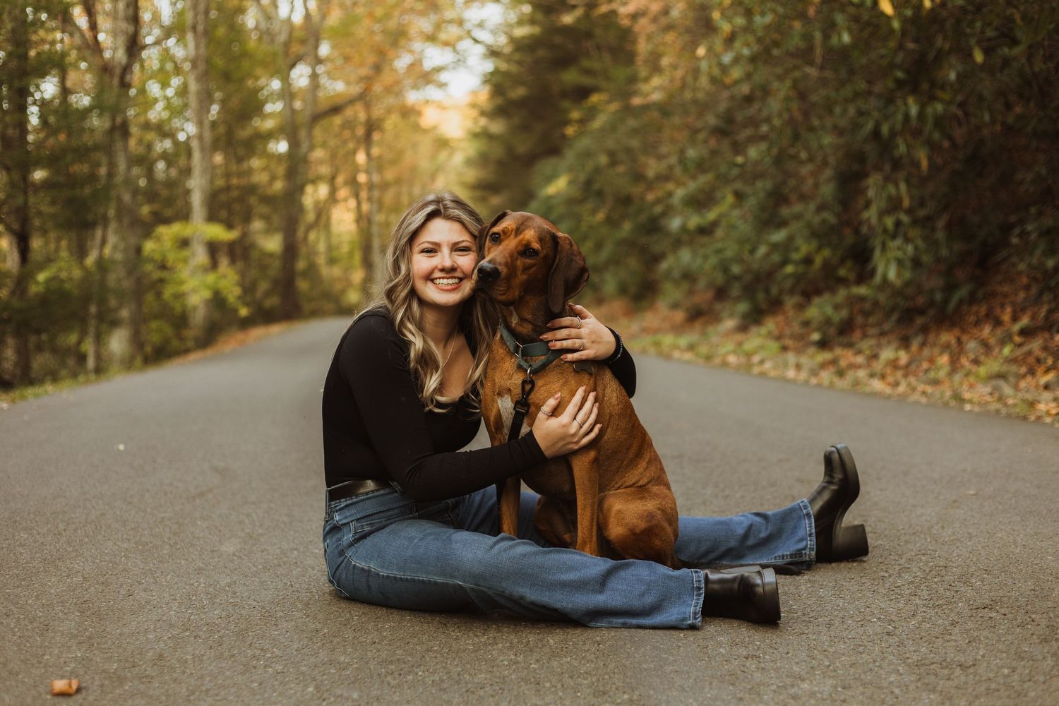 Person sitting with brown dog on paved road surrounded by fall foliage.