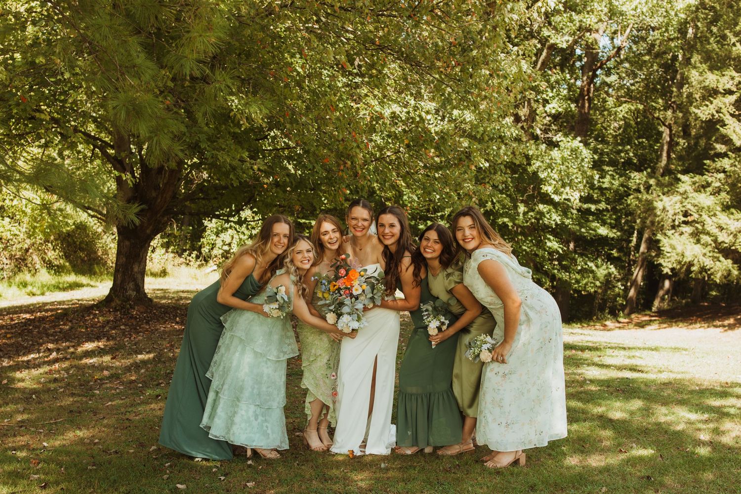 Bridesmaids in sage green dresses pose together under a large tree.
