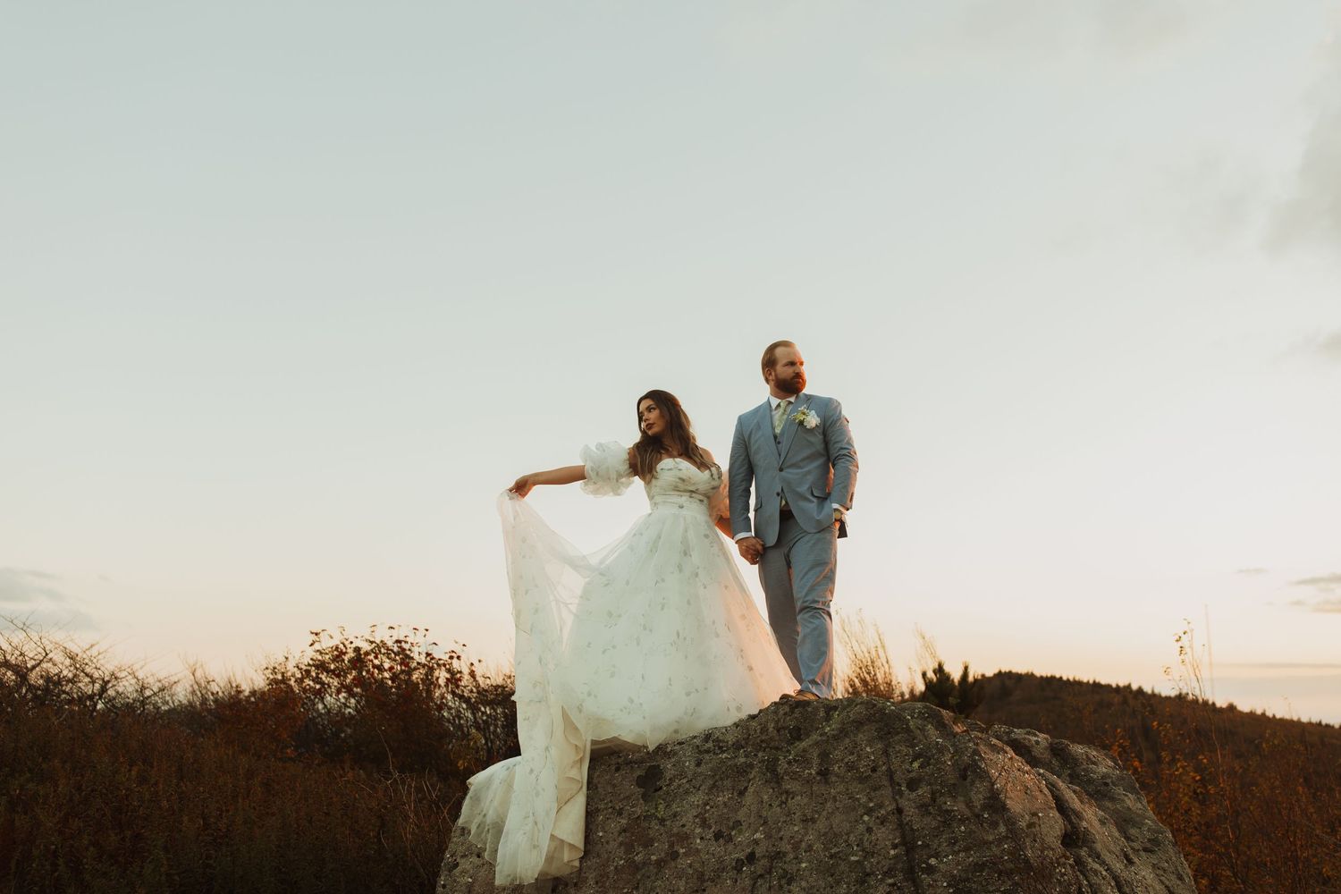 A couple stands on a rocky outcrop during sunset in wedding attire.