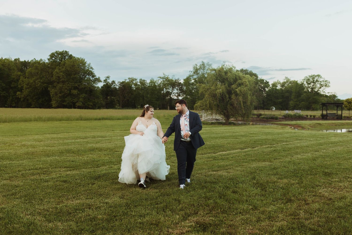 Newlyweds walk hand in hand across a scenic grassy field.