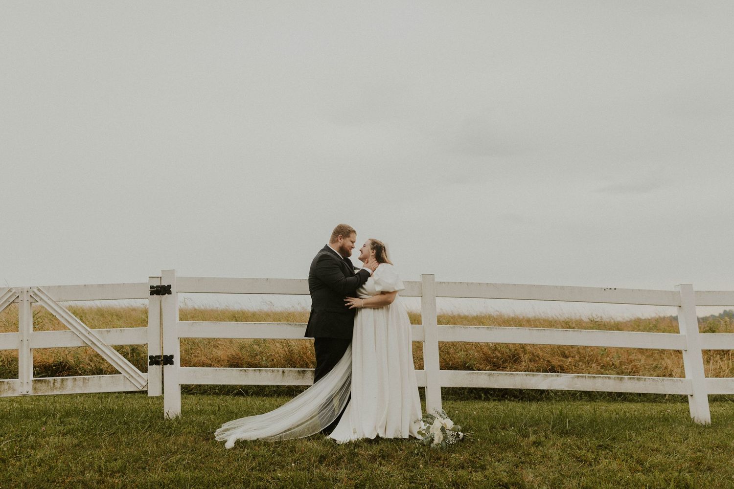 A romantic embrace by a white fence with flowing wedding dress details.