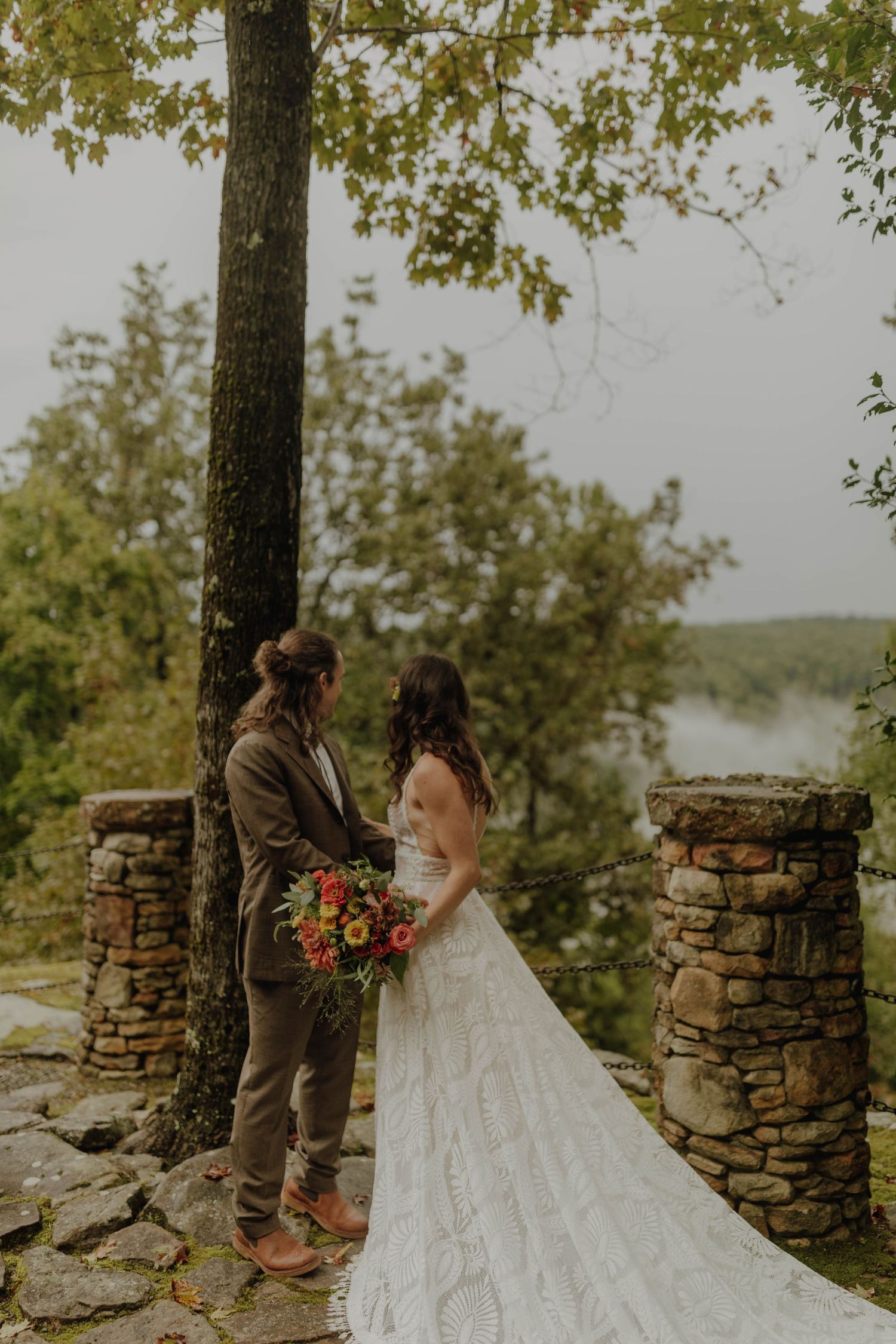 A couple stands between stone pillars during their outdoor autumn wedding ceremony.