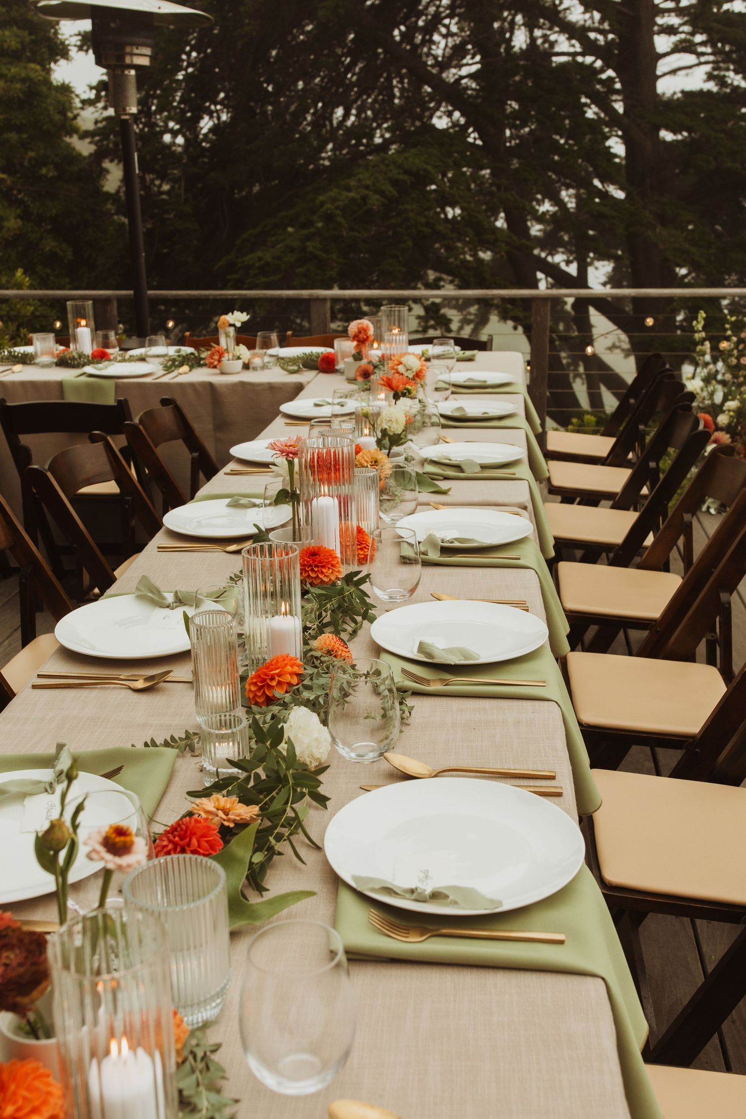 Long wedding reception table set with orange floral centerpieces and elegant place settings outdoors.
