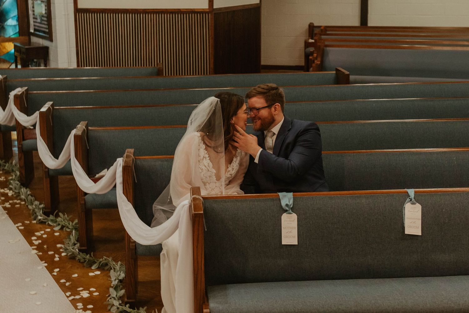 Couple sharing an intimate moment in a church pew decorated with white fabric and flower petals.