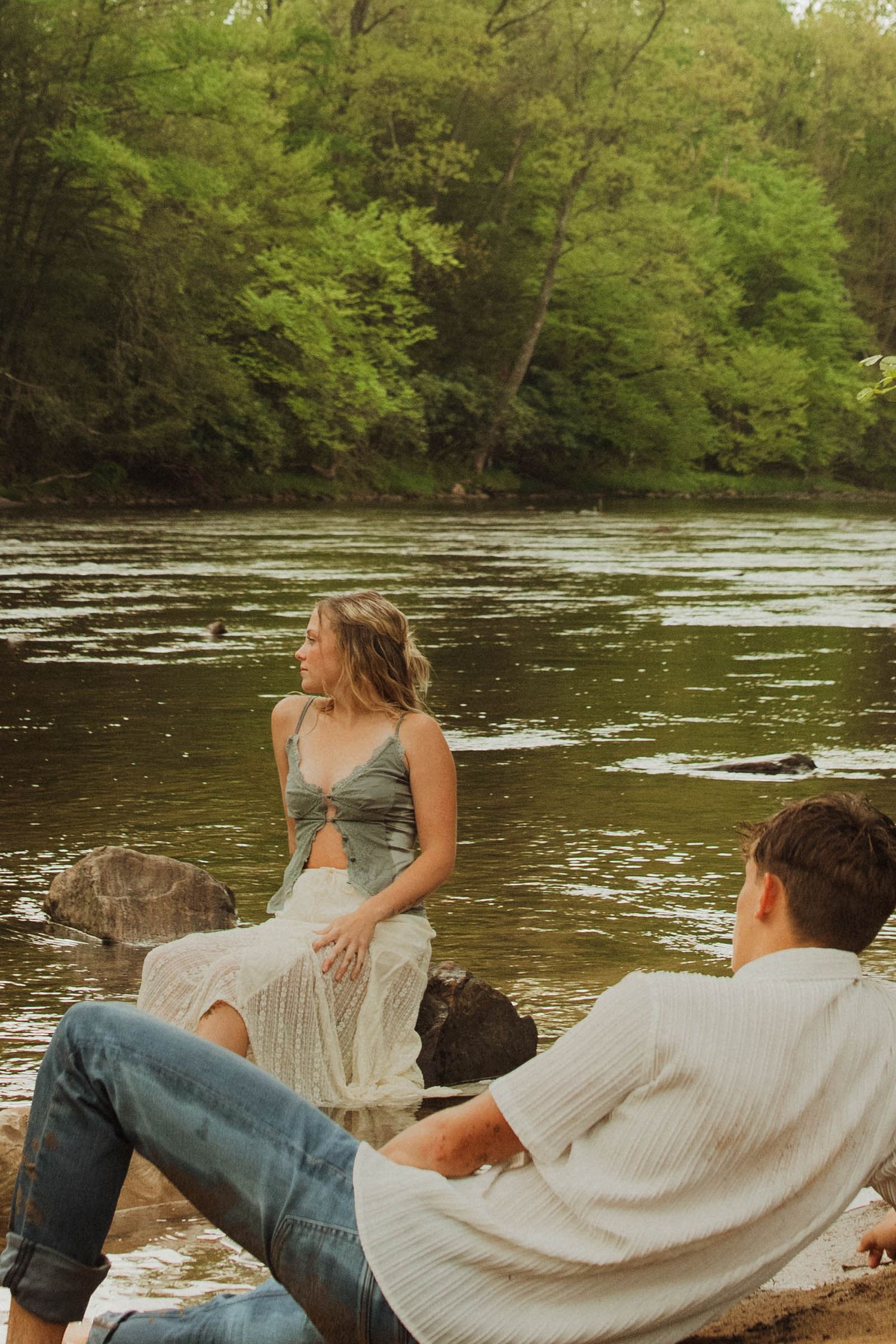 A couple relaxes on rocky riverbank surrounded by lush forest scenery.