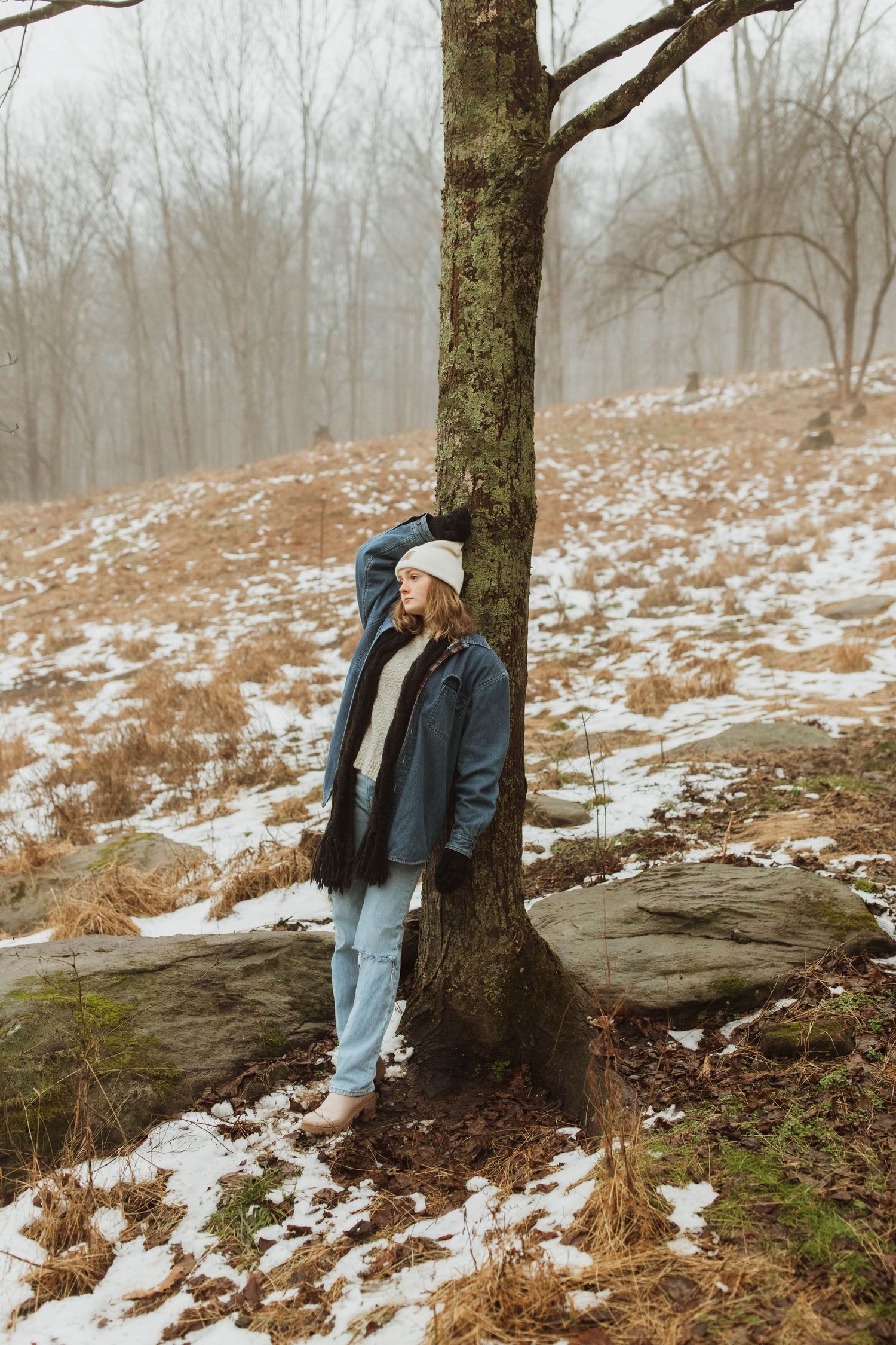 Winter scene of someone leaning against a tree trunk in snow covered woods wearing a blue coat.