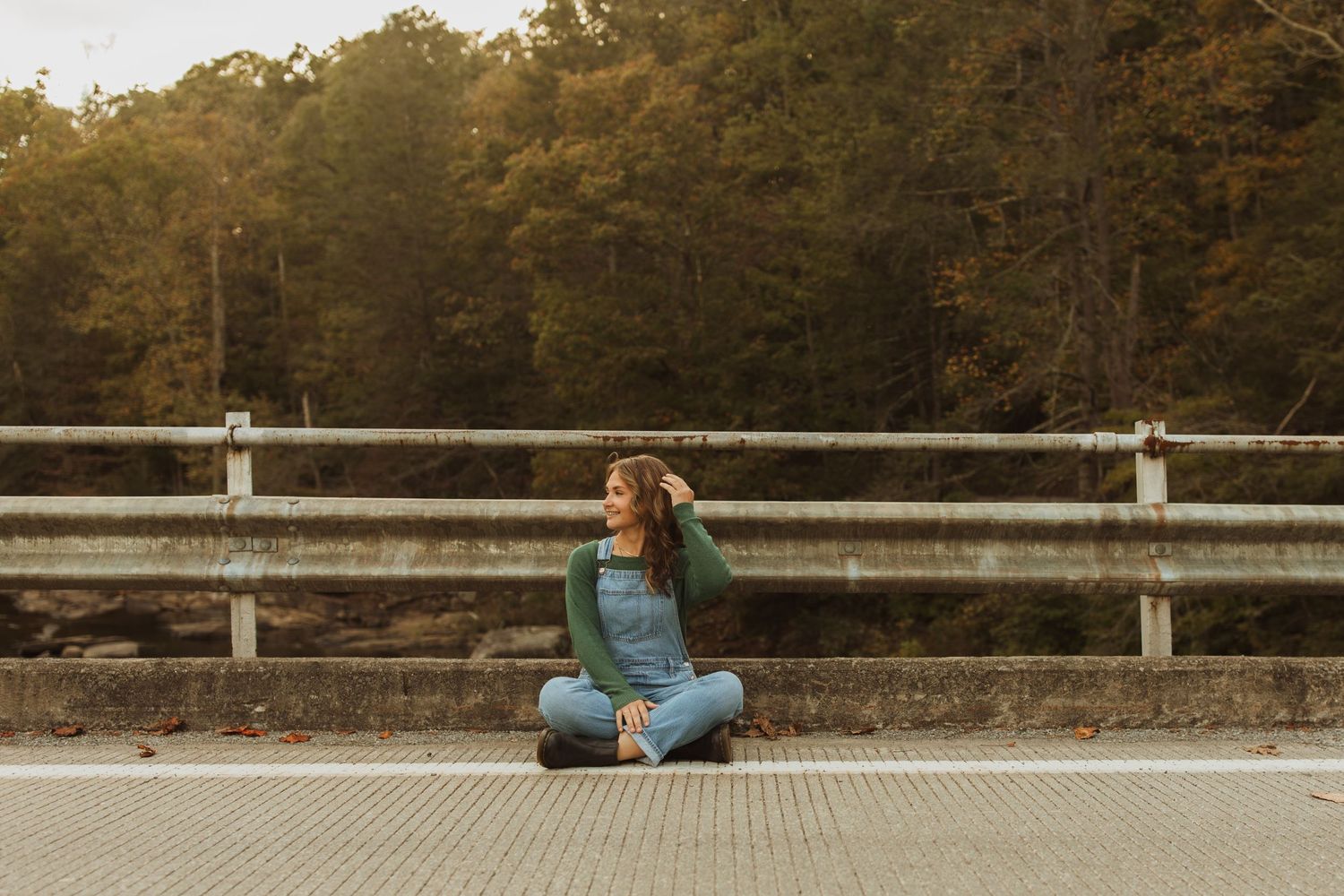 Relaxed sitting pose on bridge wearing denim overalls with forest backdrop.