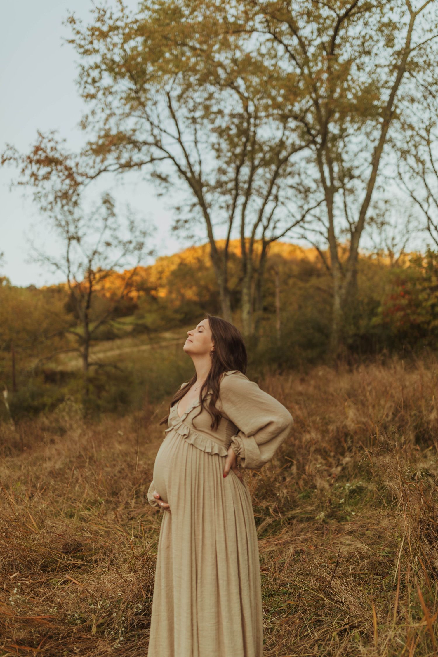 A maternity photo in a cream dress against autumn trees and rolling hills at golden hour.