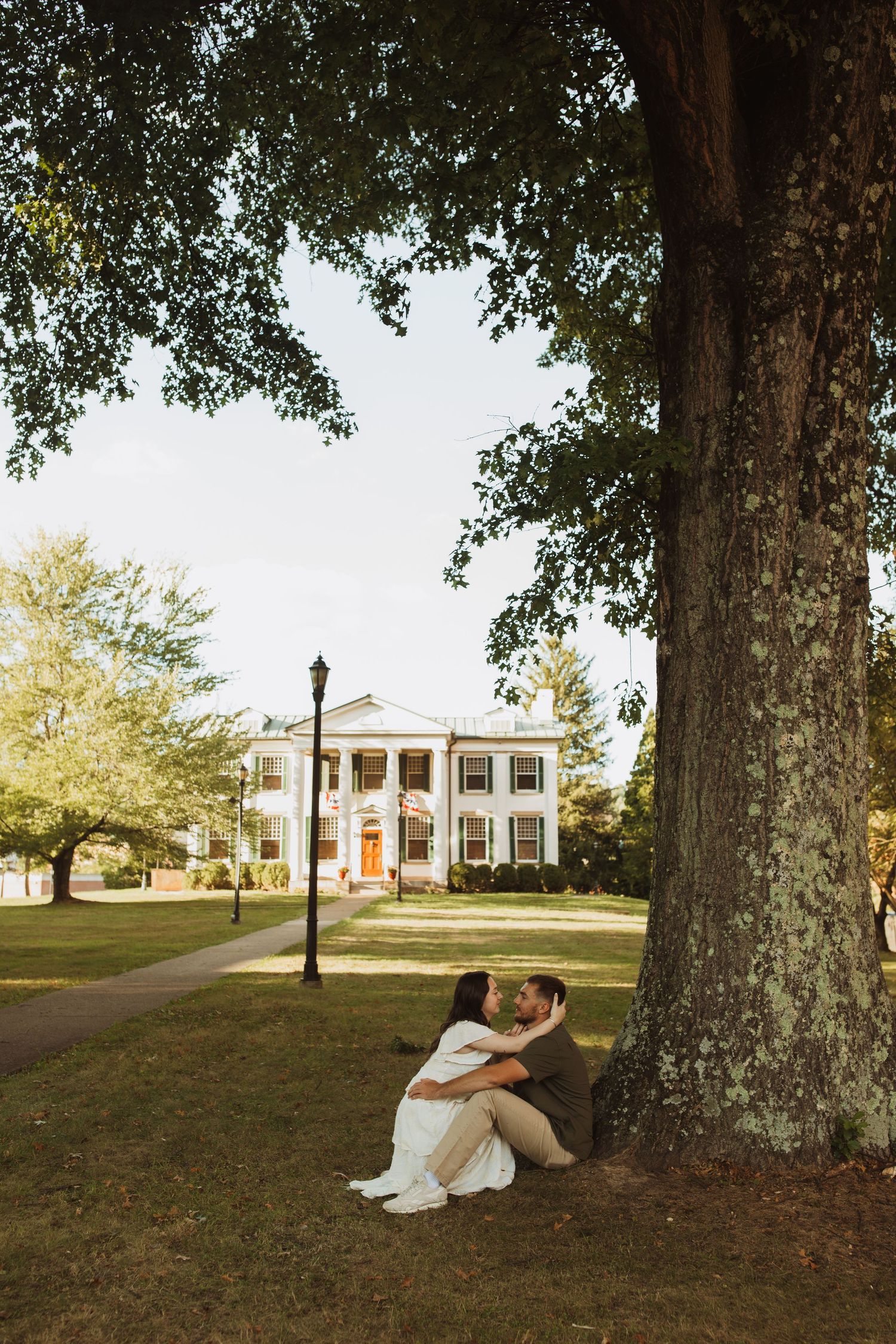 Couple sits beneath a large tree in front of a stately white columned mansion on a manicured lawn.