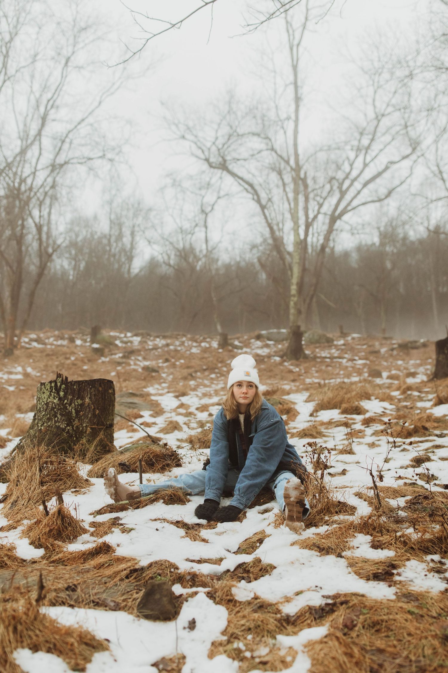 Person in blue coat and white beanie sitting in snowy woods.