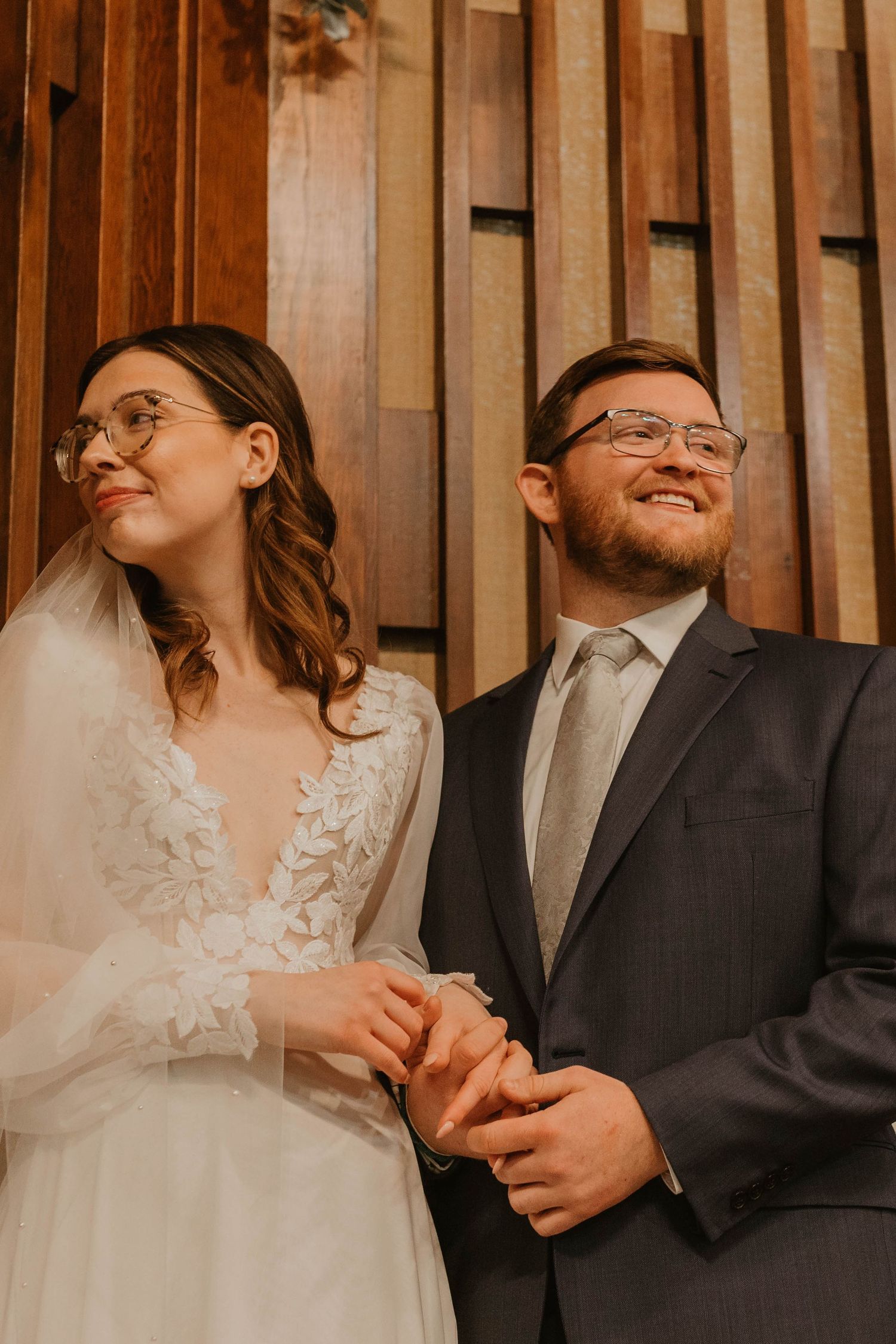 Happy couple in wedding attire standing against wooden paneled wall.