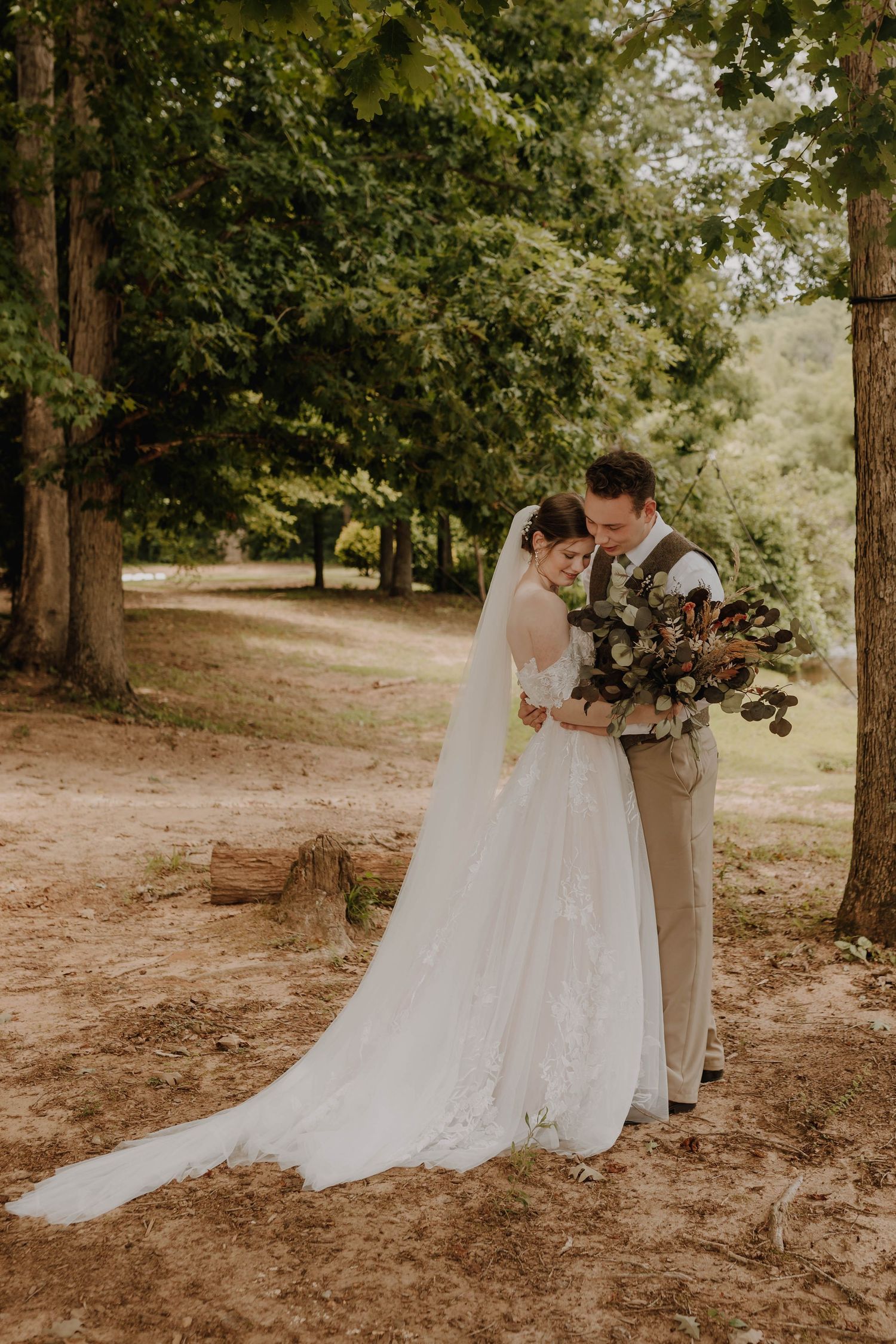 Wedding couple sharing intimate moment among trees with beautiful bouquet.