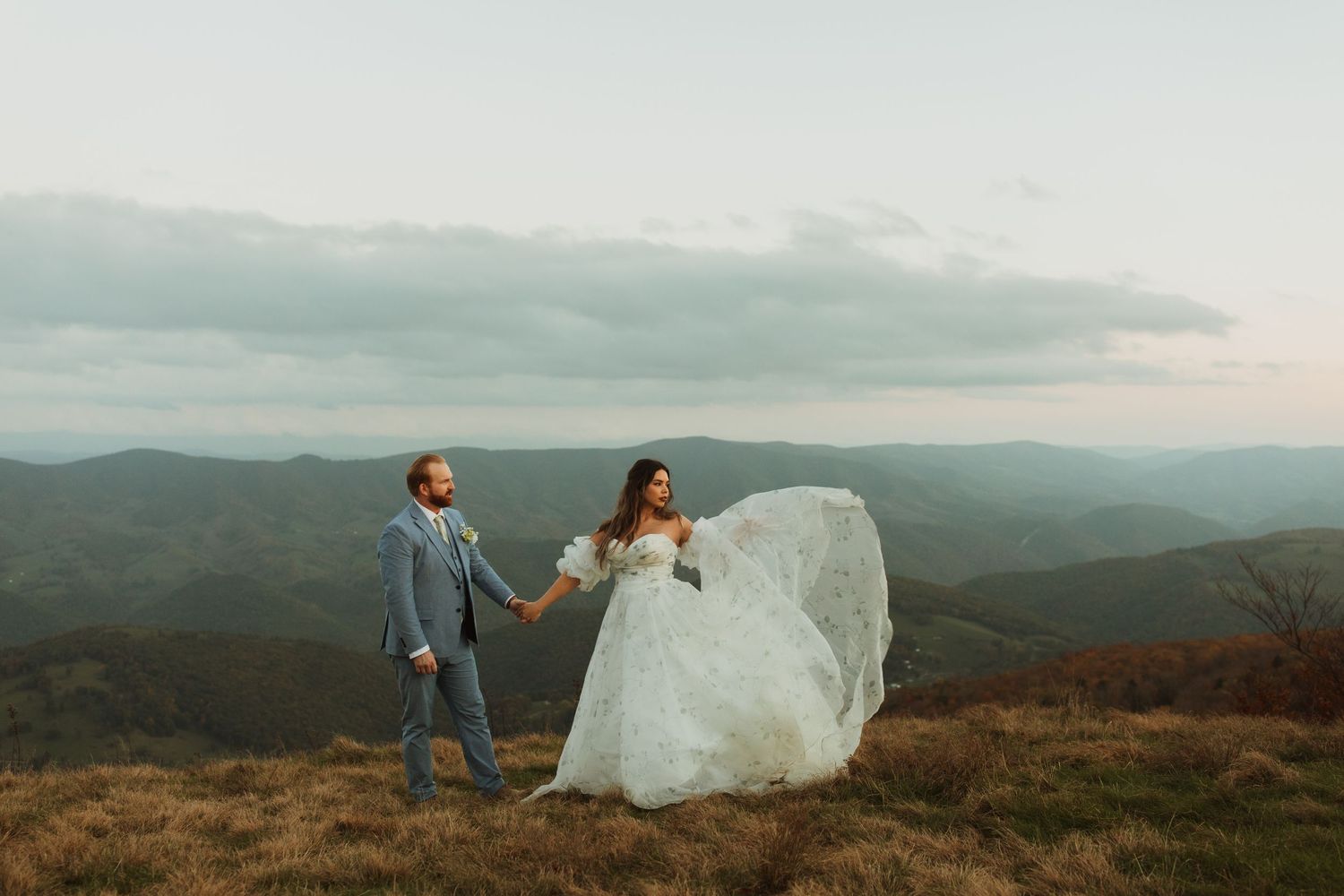 Wedding couple poses on mountain summit as bride's dress flows in the wind.